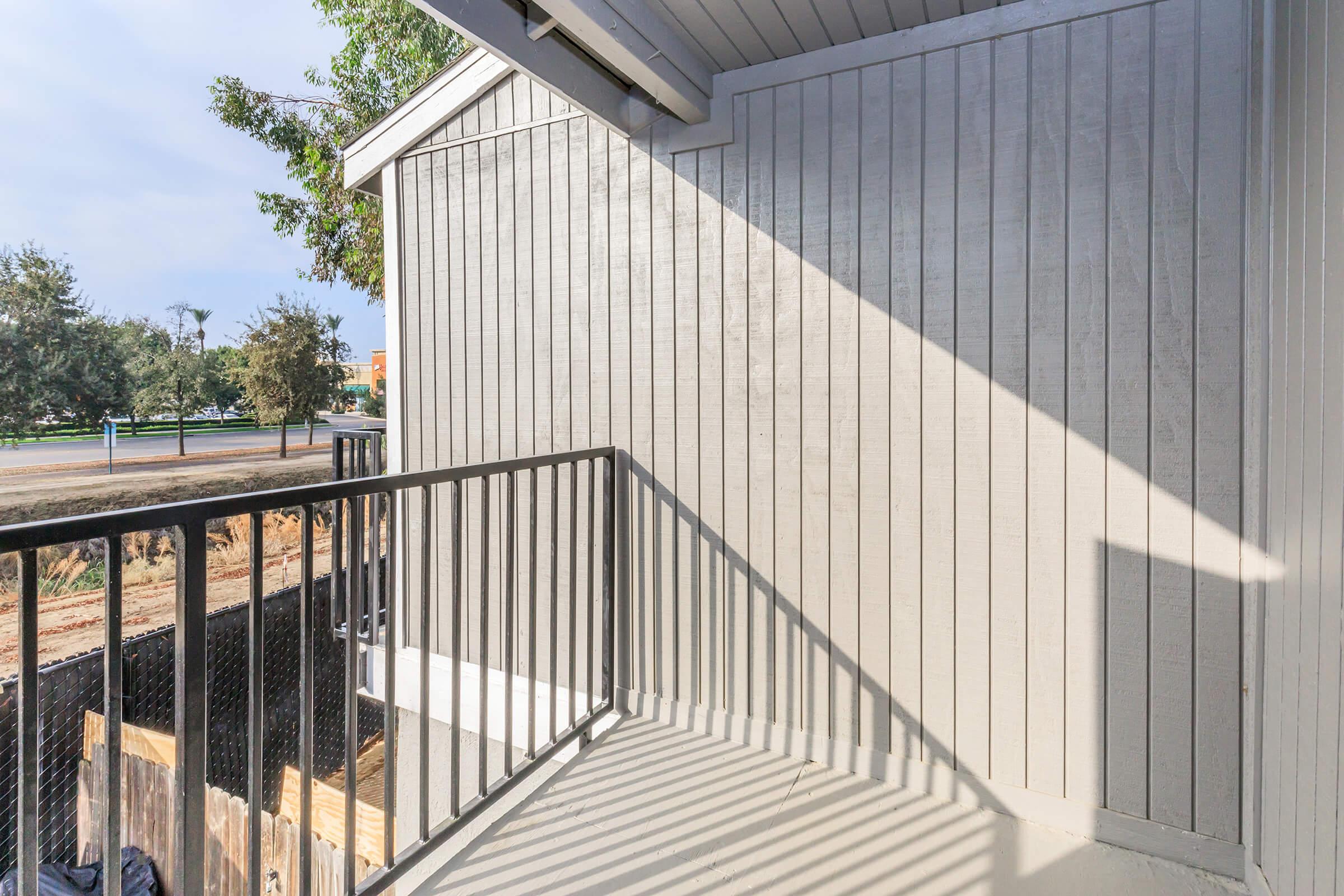 A corner balcony with gray wooden siding and a black railing. Sunlight casts shadows on the floor, and greenery is visible in the background along with a road and construction area. The scene conveys a sense of quiet outdoor space.