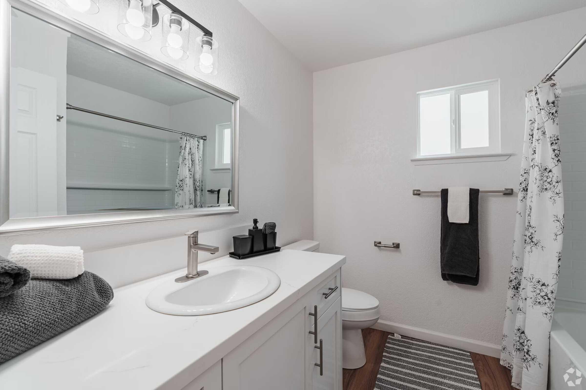 A modern bathroom featuring a white vanity with a sink, a large mirror, and stylish lighting. The decor includes a gray bath mat, neatly rolled towels, and a black decorative container. There’s a window allowing natural light, and a shower area is visible with a white curtain.