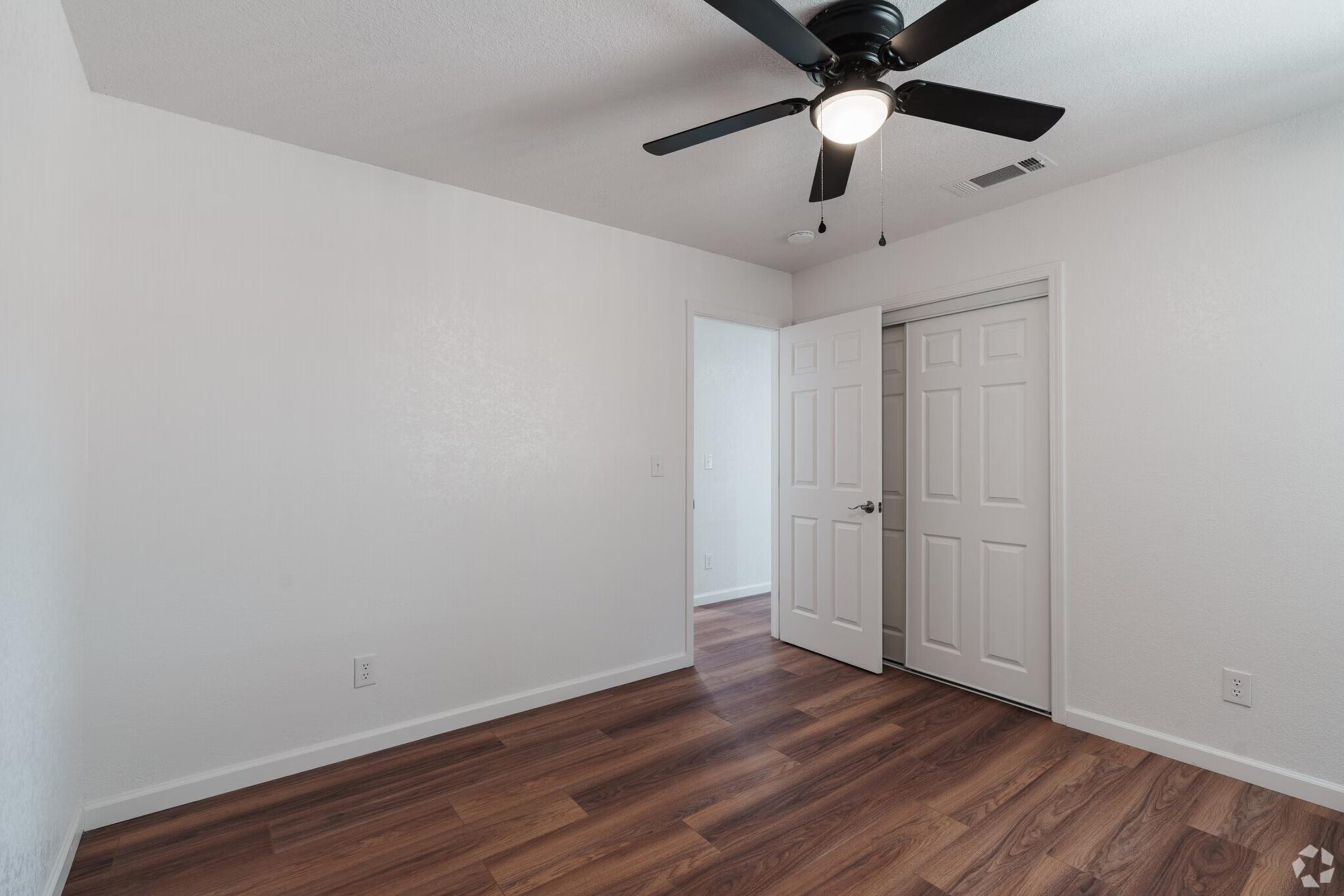 Empty room with white walls, a ceiling fan, and a door leading to another room. The floor is wooden, and there's a closet door visible. The room has ample natural light, making it appear bright and spacious.