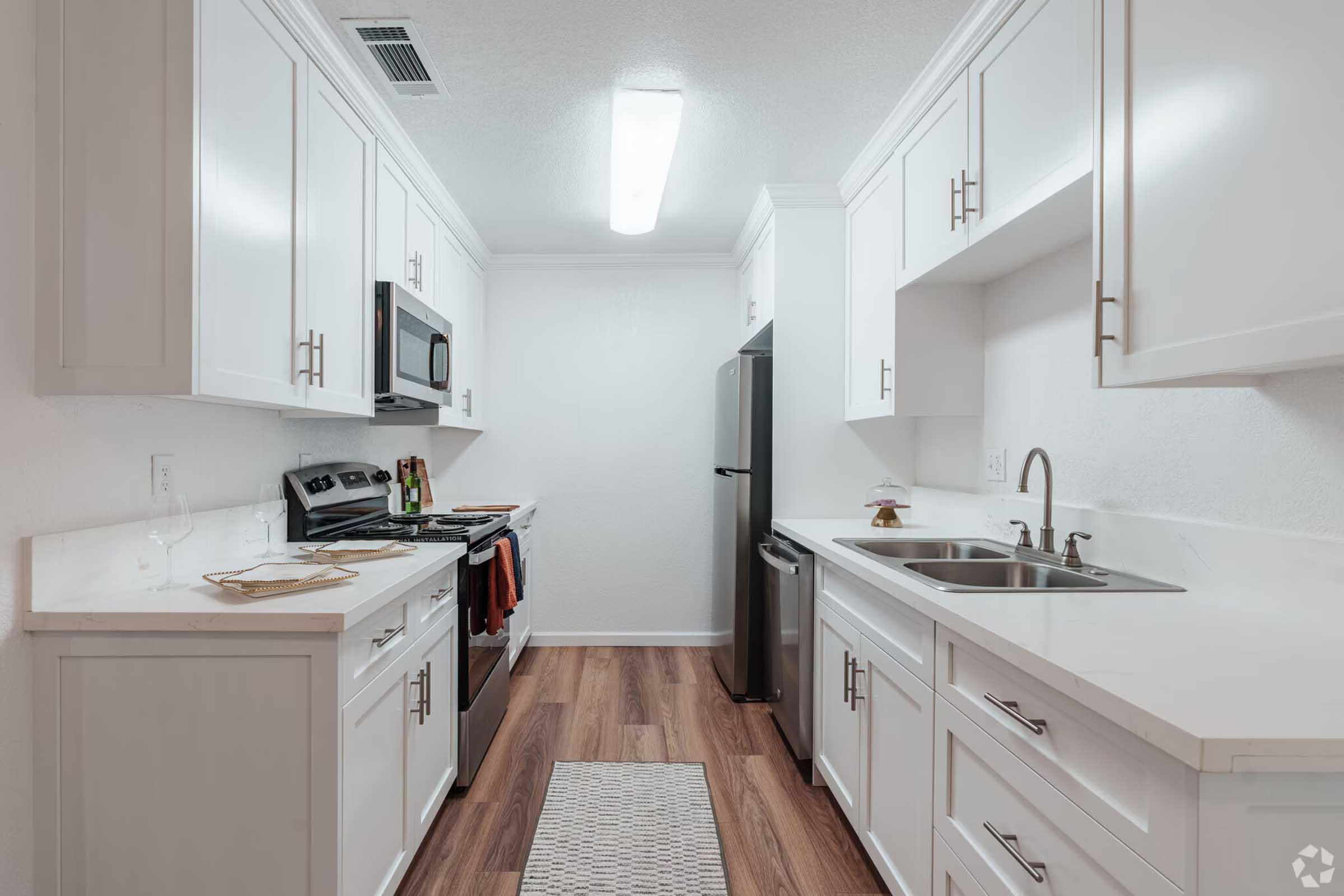 Modern kitchen featuring white cabinets, stainless steel appliances including a microwave, oven, and refrigerator. The countertop is white with a light wood floor, and a small rug is placed in the center. The space is well-lit with overhead lighting.