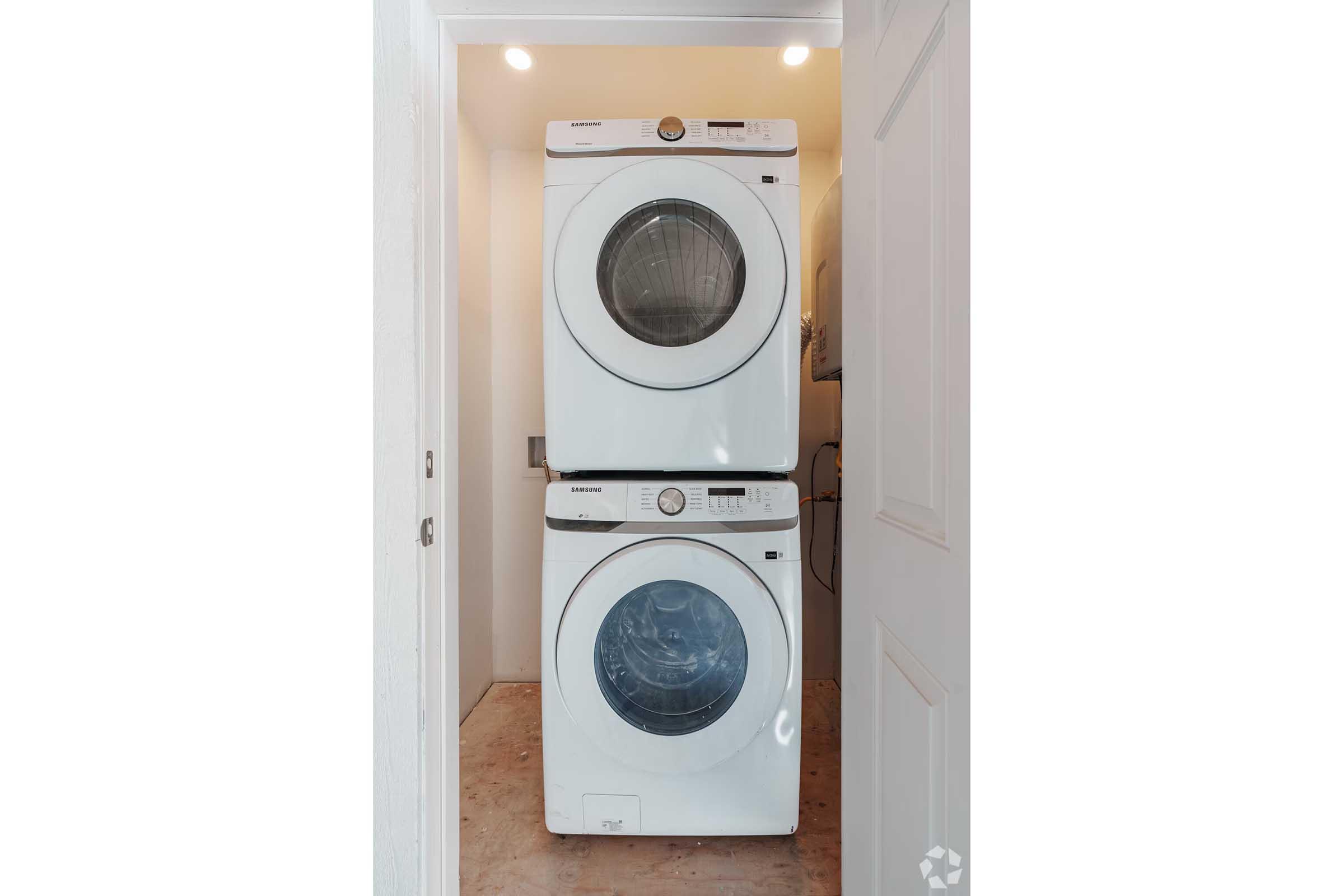 A stackable washer and dryer unit positioned against a wall in a small laundry space. The appliances are white with control panels visible, and natural light is coming from above, highlighting the clean and organized appearance of the space.