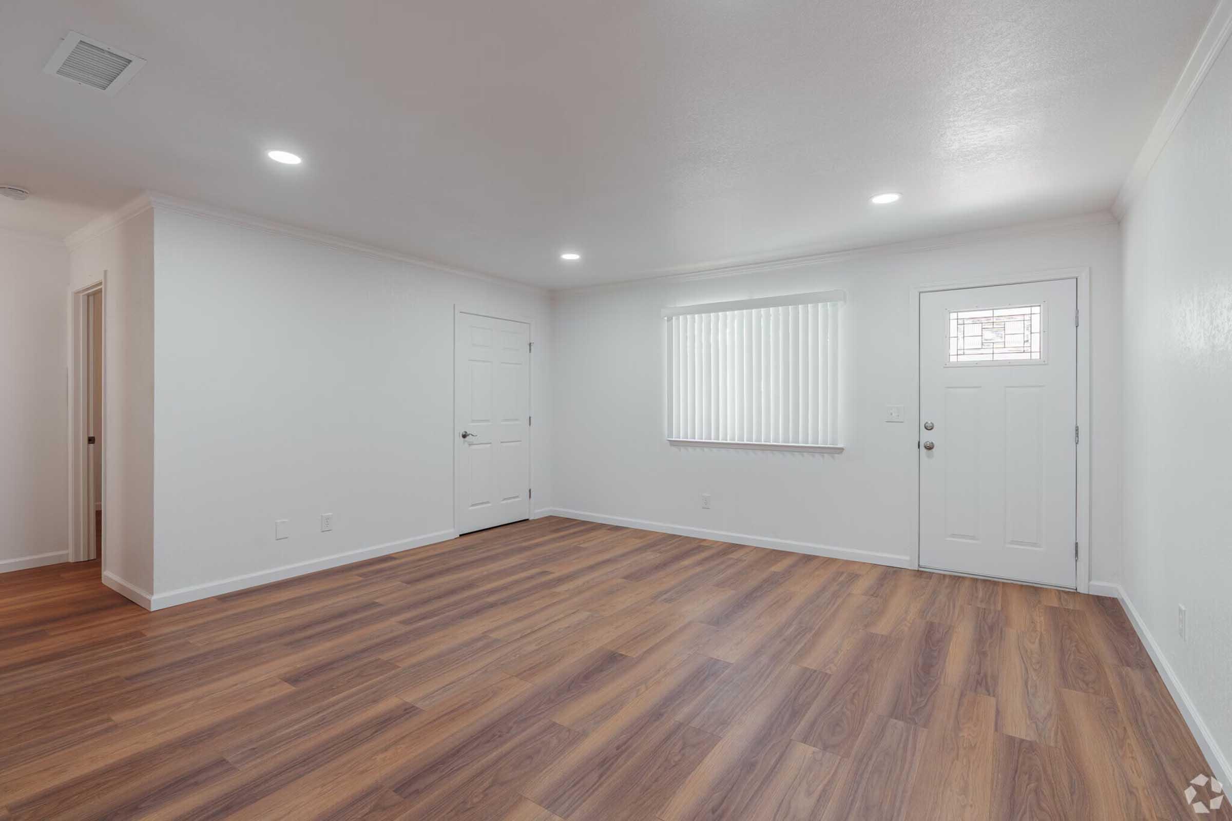 A spacious, well-lit room featuring light-colored walls, hardwood flooring, and a front door with a glass insert. There are two closed doors leading to separate areas and a window with vertical blinds allowing natural light to fill the space.