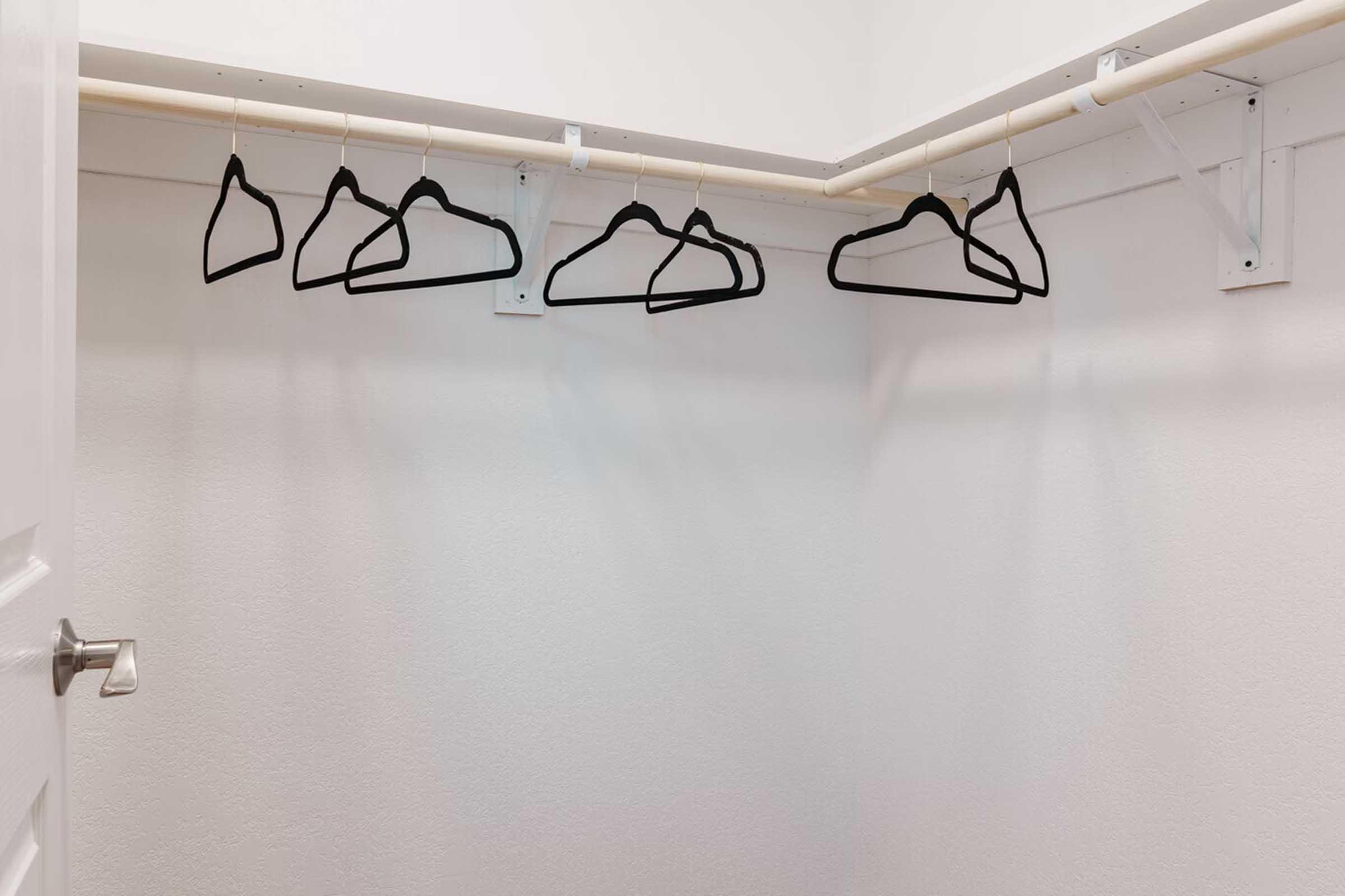A neatly organized closet with a white wall and a hanging rod. Several black hangers are suspended from the rod, indicating storage space for clothing. The floor is visible but empty, giving a clean and minimalistic appearance.