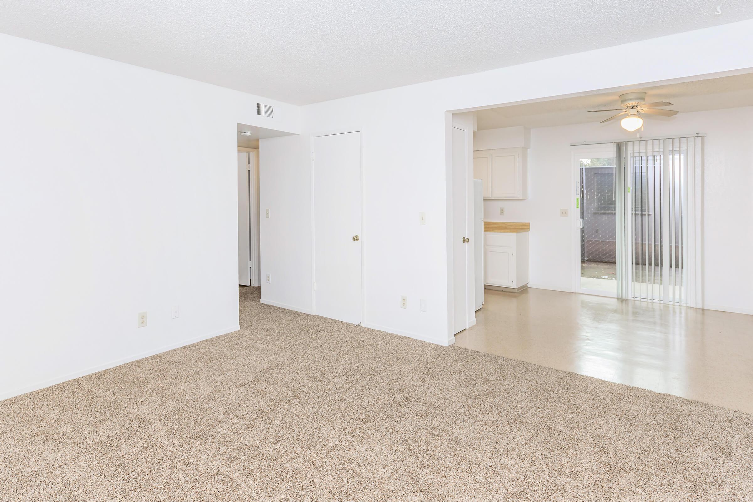 A bright, spacious living area with light beige carpet and white walls. There is a doorway leading to another room on the left, and an open kitchen area visible in the background. A sliding glass door provides natural light and leads to an outdoor space. A ceiling fan hangs above, adding to the modern feel.