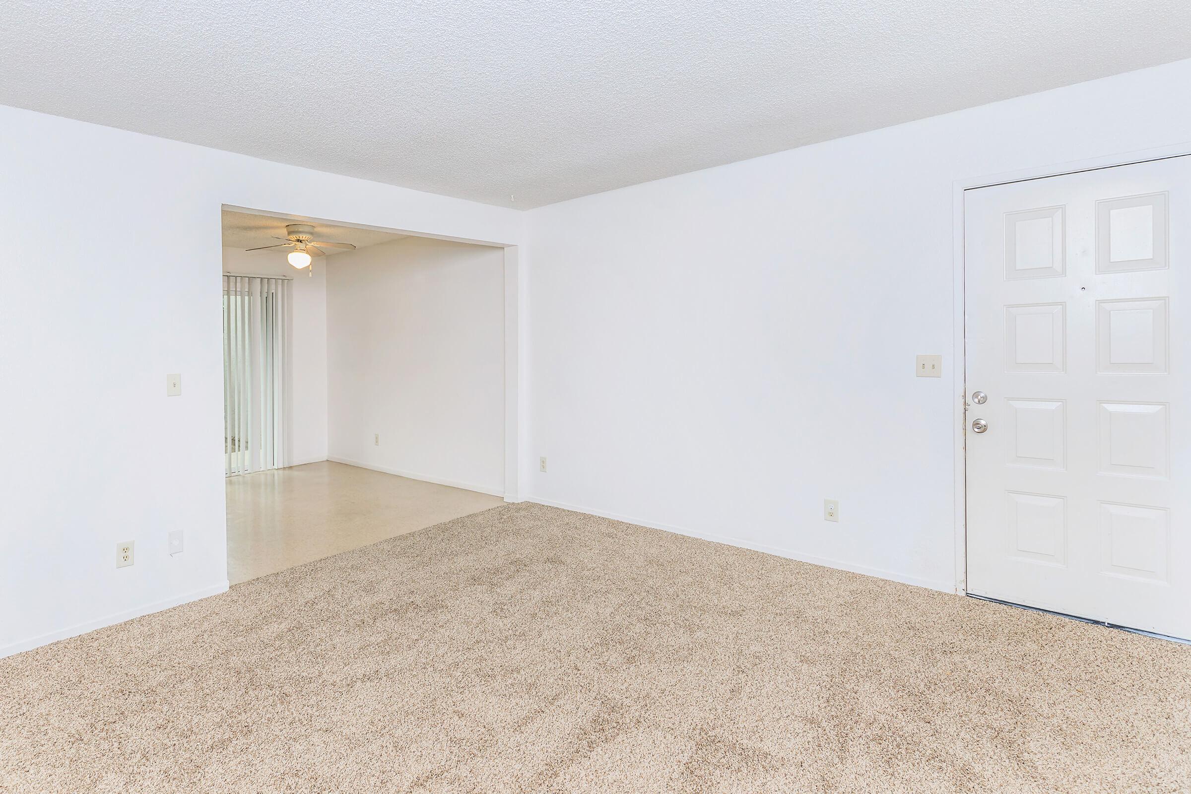 A spacious living area featuring light-colored carpet, white walls, and an entrance door. A doorway leads to another room with a ceiling fan, while large sliding glass doors are visible in the adjacent space, allowing natural light to brighten the area.