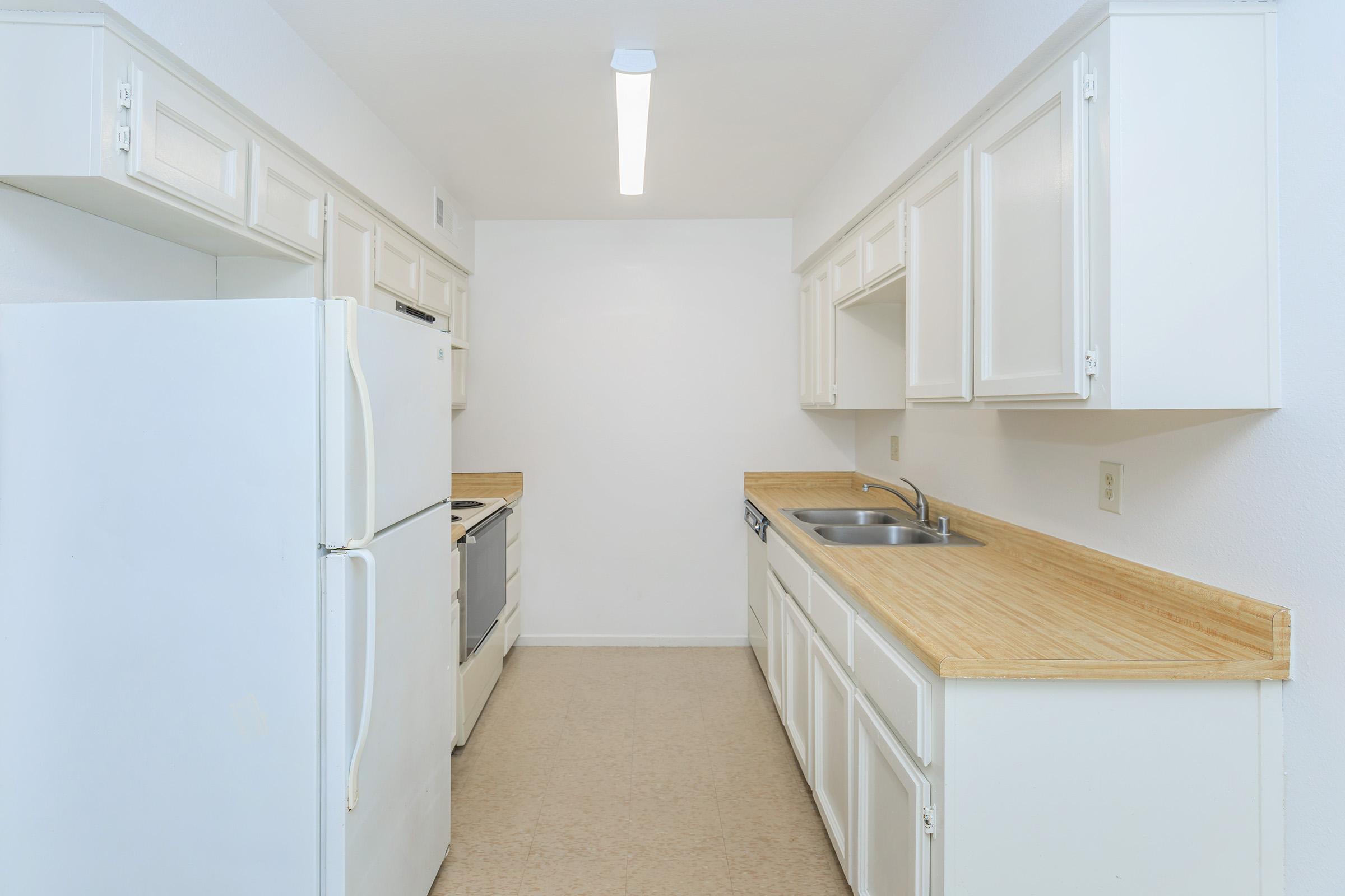 A clean, modern kitchen with white cabinetry and a light wood countertop. The space includes a refrigerator, oven, and sink, with a large window providing natural light. The walls are painted white, and the flooring is a light beige. A bright overhead light fixture illuminates the area.