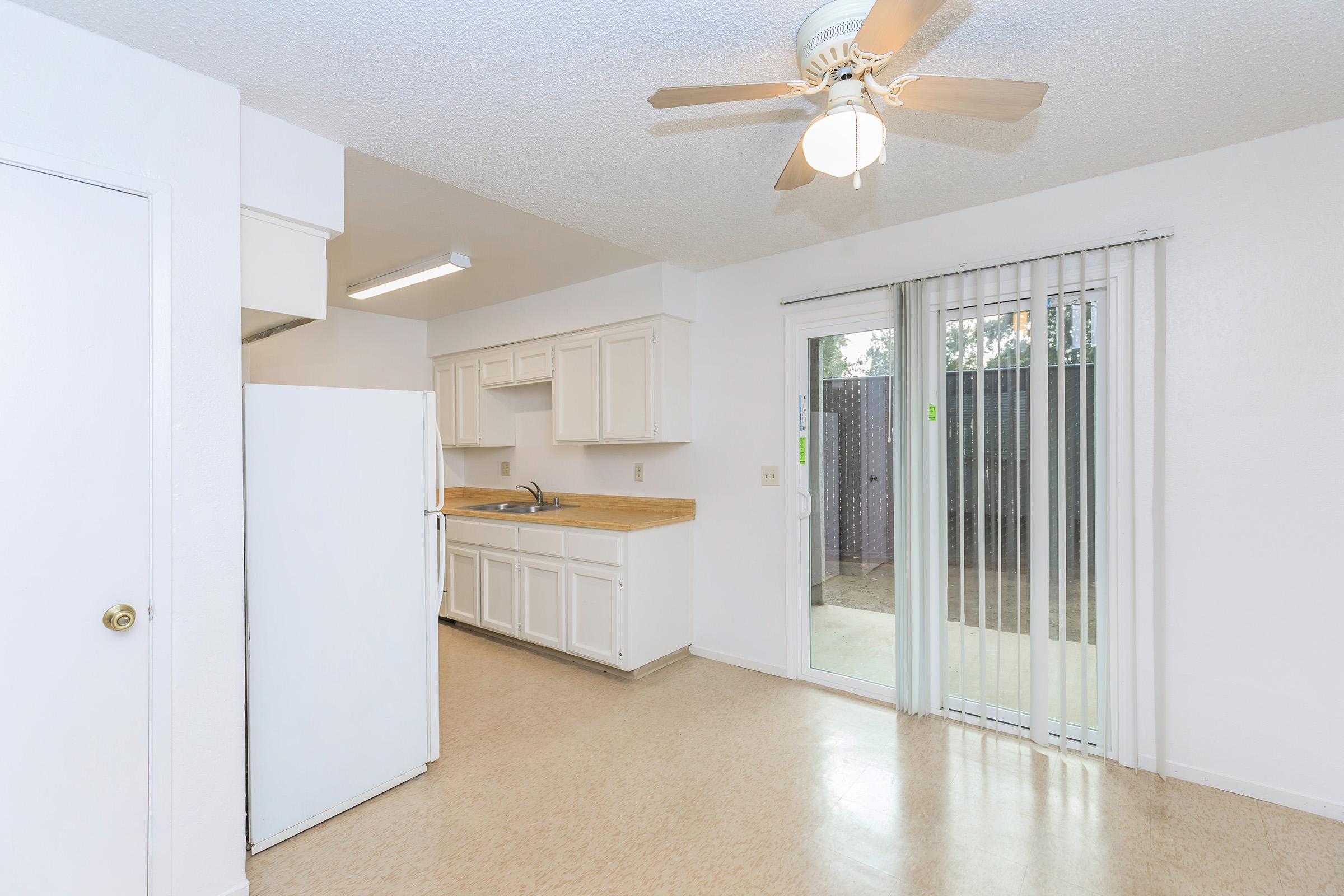 A bright kitchen featuring white cabinetry, a refrigerator, and a countertop with a sink. A ceiling fan provides ventilation, and there is a sliding glass door leading to an outdoor area. The floor is light-colored and the walls are painted white, creating a clean, open space.