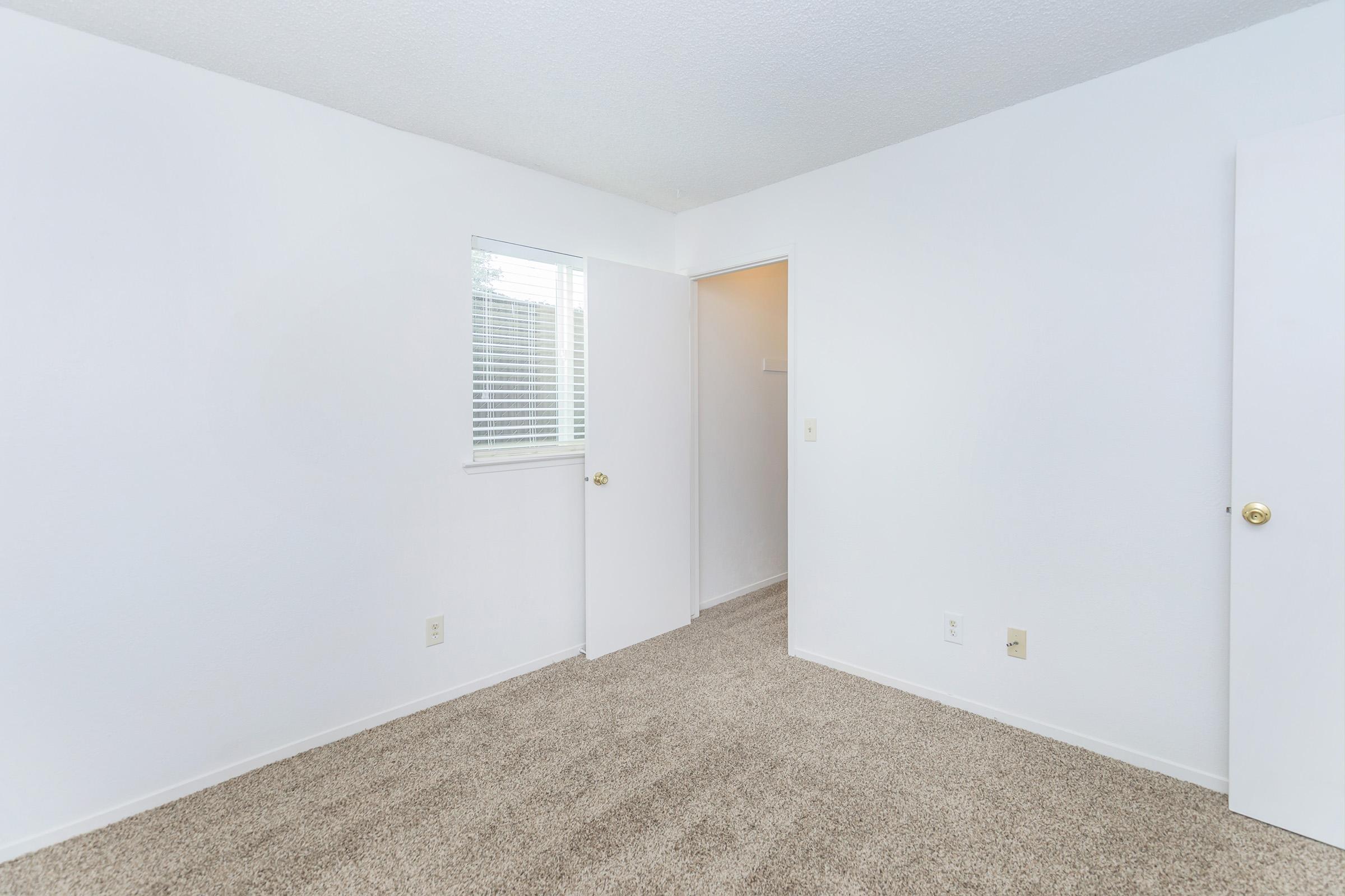 Empty room with light-colored walls and beige carpet. A window with blinds is on one wall, and a door leads to a hallway. The room appears well-lit and clean, ideal for use as a bedroom or office space.