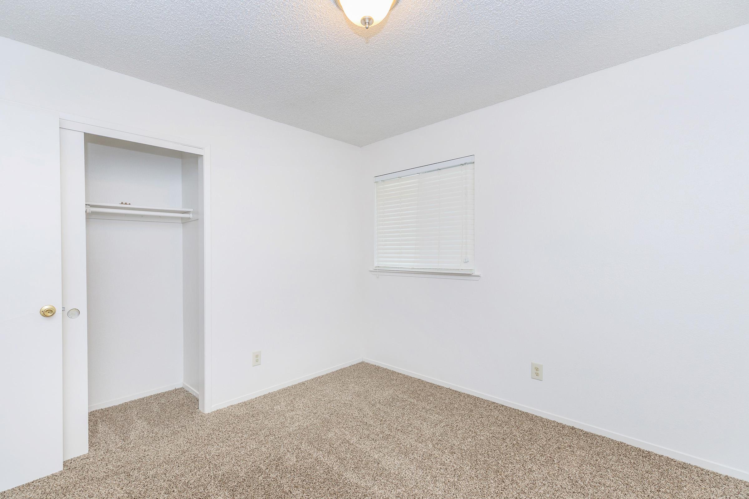 A small, empty bedroom featuring light beige carpet, white walls, and a single window with blinds. There is a built-in closet with a sliding door on the left side of the room. The ceiling has a light fixture, and the overall space appears clean and well-lit.