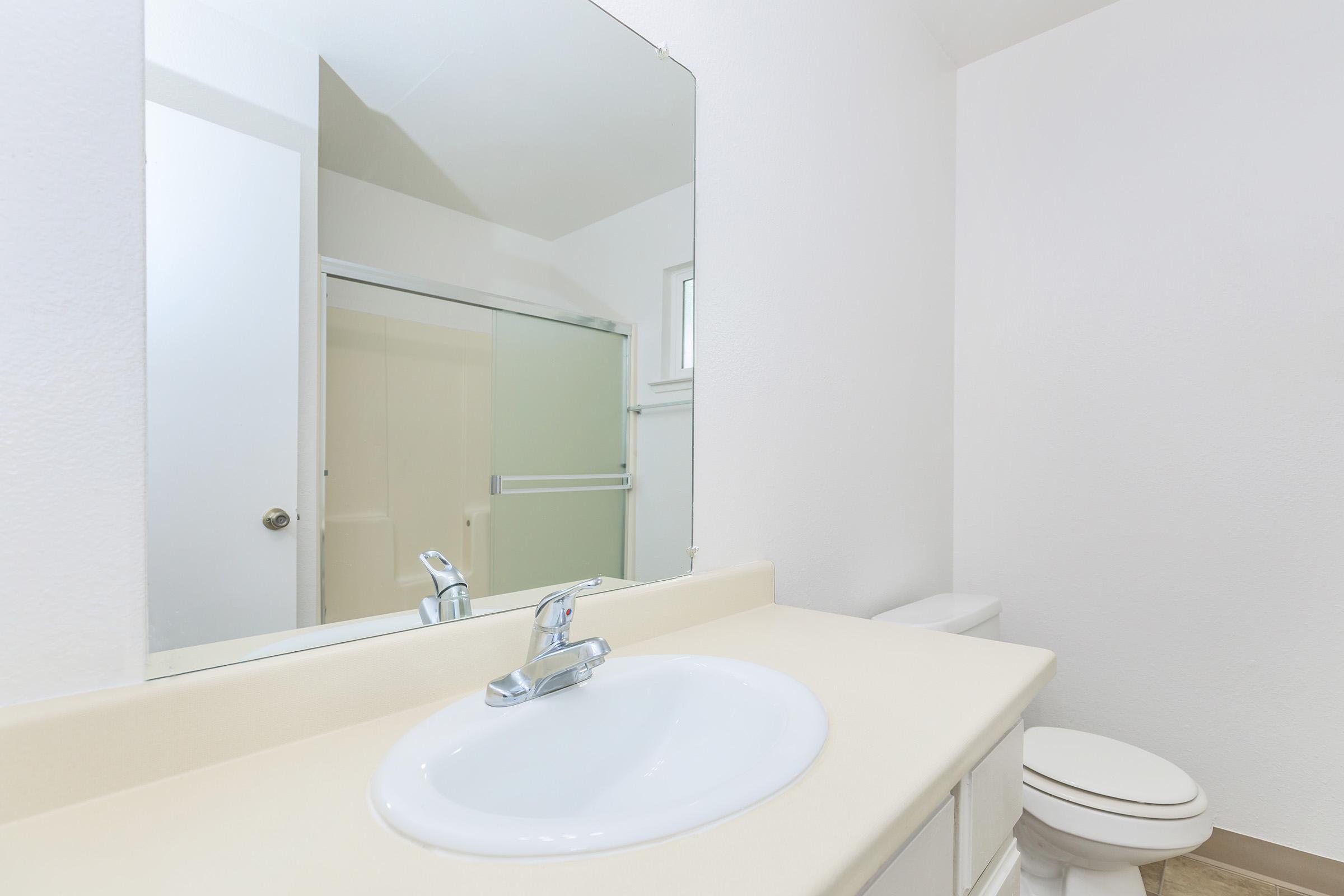 A clean and modern bathroom featuring a white countertop with a sink and faucet, a large mirror above the sink, a toilet nearby, and a glass shower enclosure in the background. The walls are painted white, creating a bright and spacious feel.