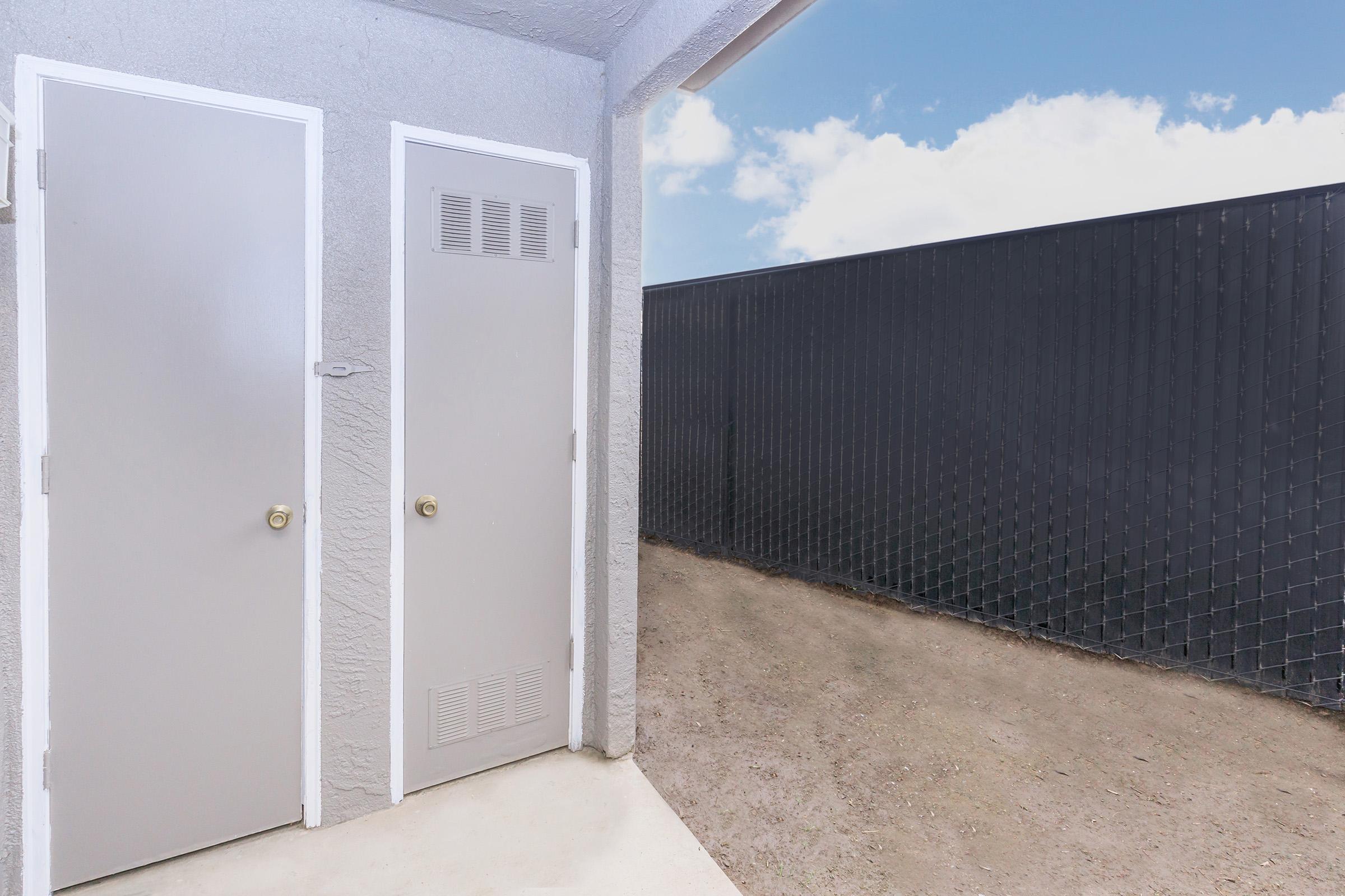 A view of a gray exterior featuring two doors on the left, with one partially open. To the right, a fenced area with a chain-link fence stretches towards the horizon under a blue sky with scattered clouds. The ground is bare and unpaved.