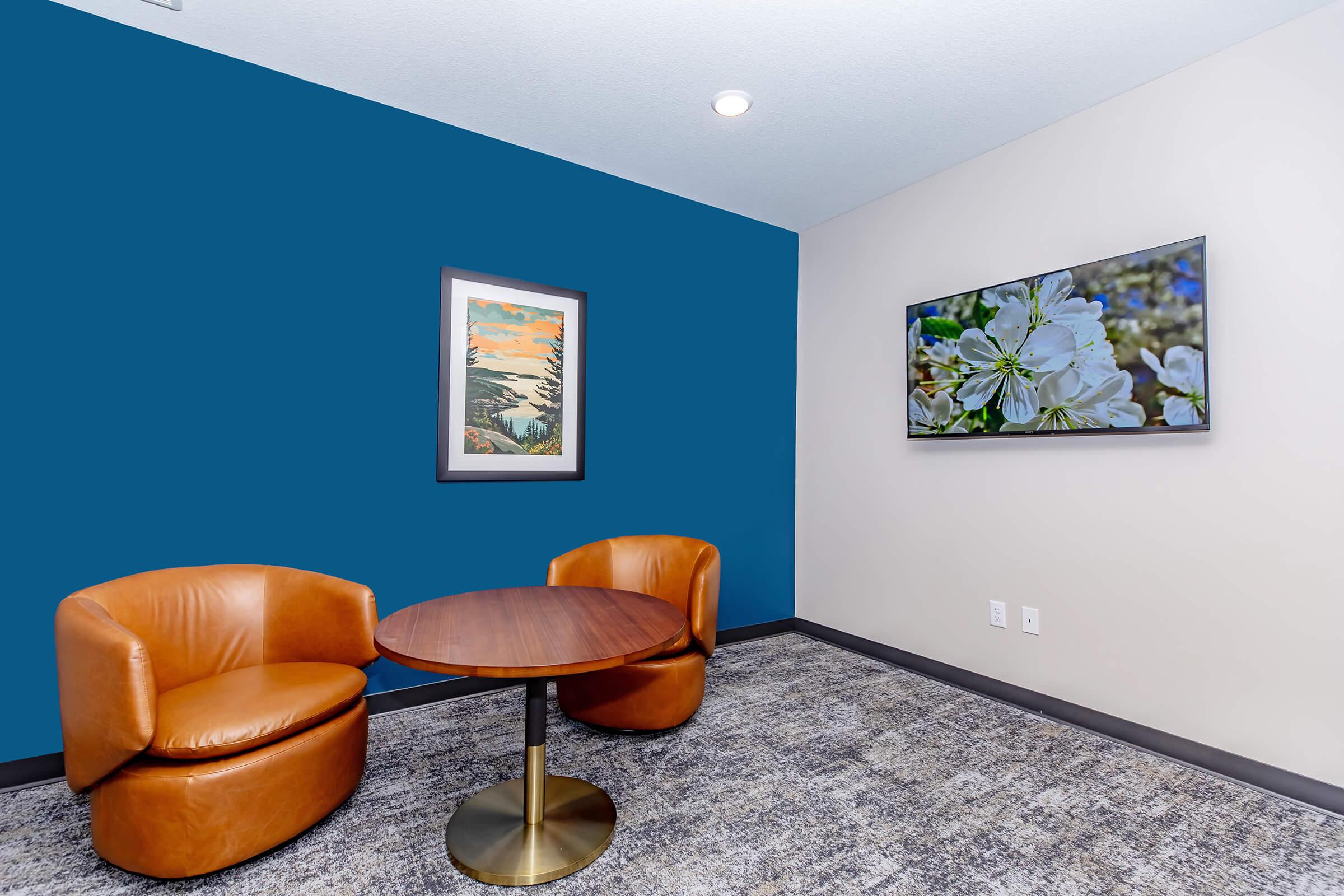 A cozy seating area featuring two brown leather chairs and a round wooden table on a textured gray carpet. The wall behind the chairs is painted in a vibrant blue, while one wall displays a framed landscape painting and another showcases a flat-screen TV with floral imagery.