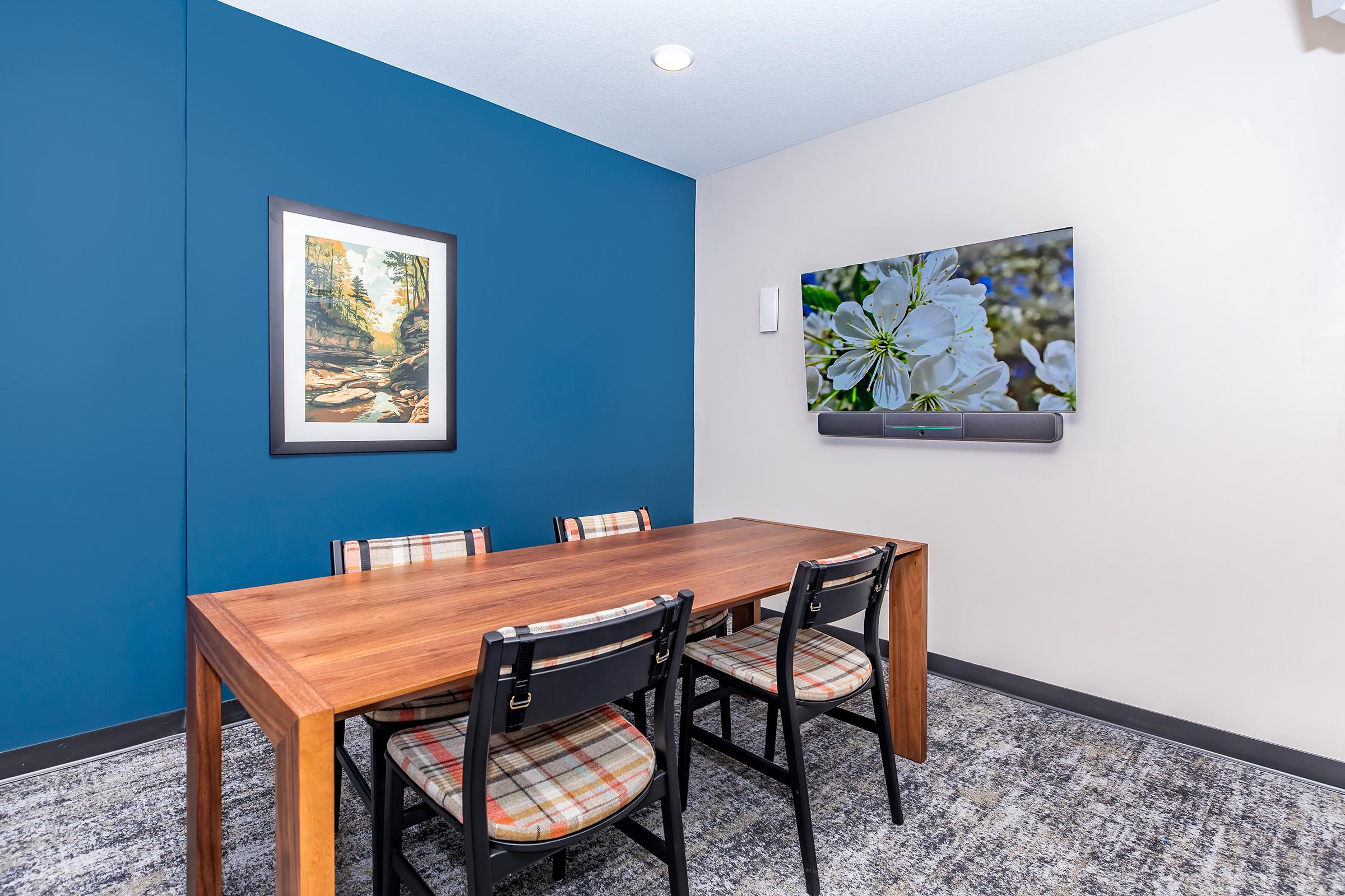 A modern meeting room featuring a wooden table with four plaid chairs. One wall is painted blue, while another has a framed landscape painting. A flat-screen TV mounted on the wall displays a floral image. The flooring is carpeted in neutral tones.