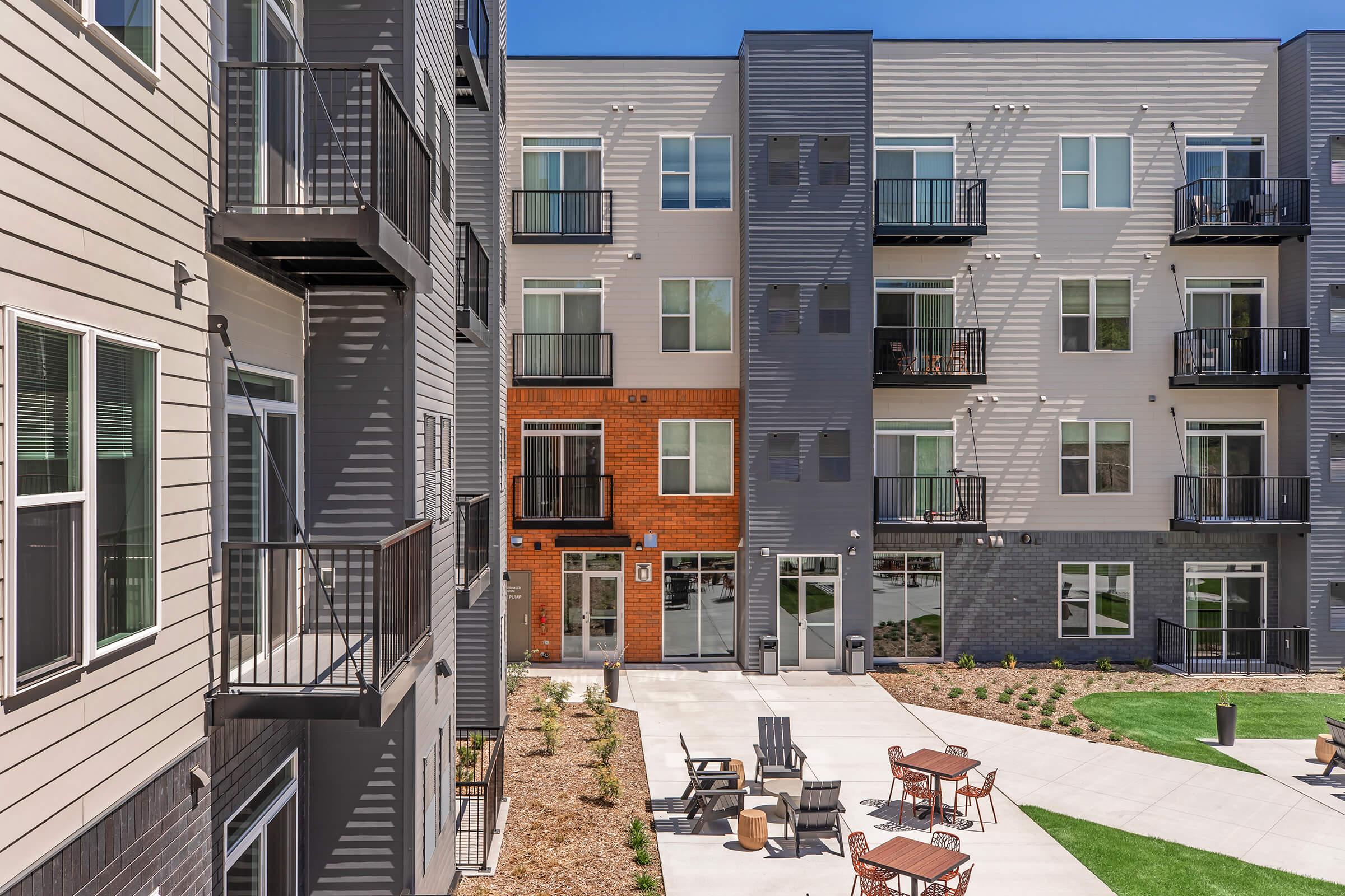 Modern apartment complex featuring multiple stories, with a mix of gray and brick facades. Balconies are visible on the upper floors, and the ground-level area includes a patio with seating and landscaped green space. Bright blue sky overhead adds to the vibrant atmosphere.