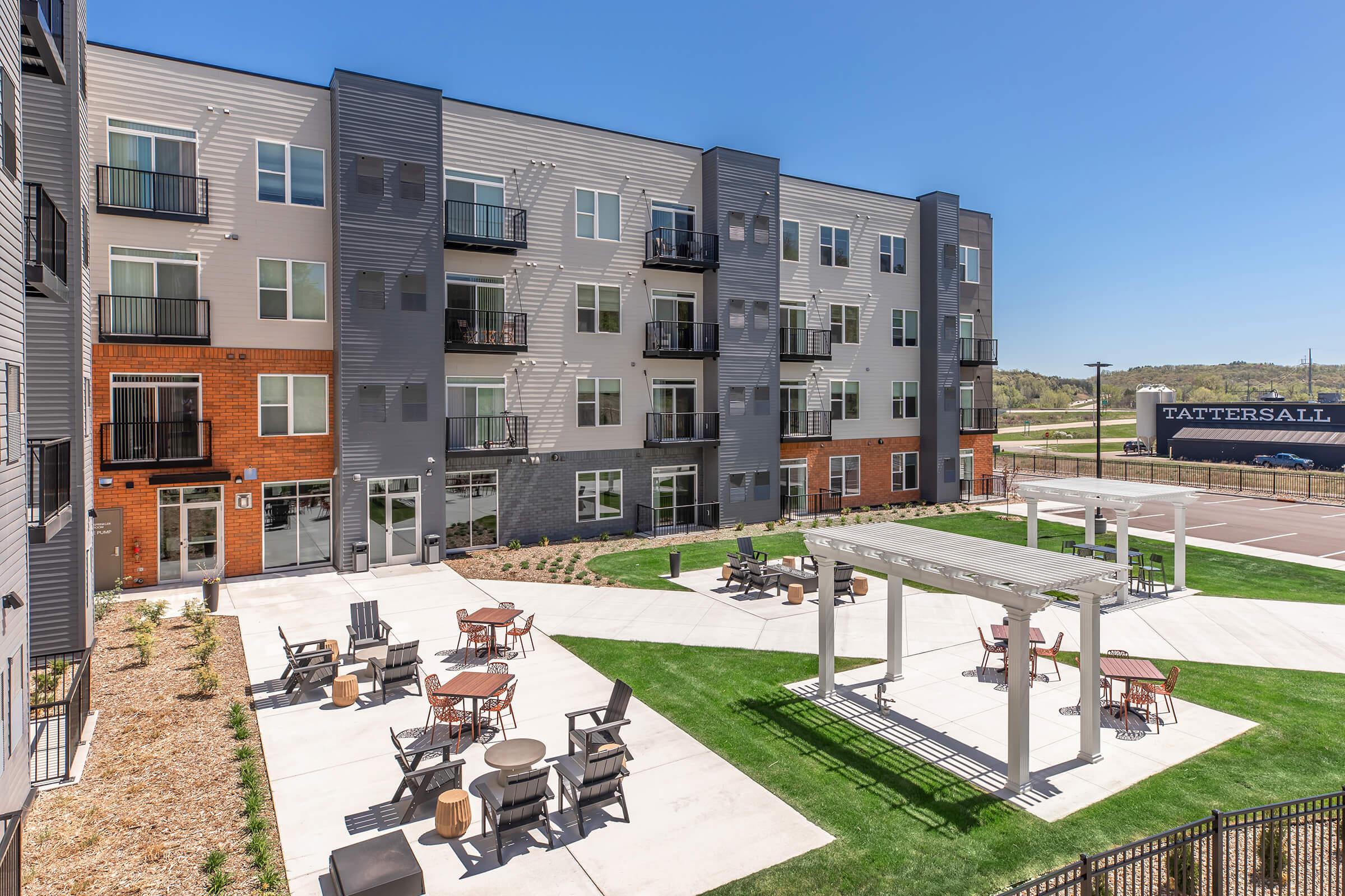 Modern multi-story apartment building with a mix of brick and siding exteriors. The ground level features a landscaped outdoor area with seating, fire pits, and pergolas. Clear blue skies and greenery surround the building, creating a welcoming atmosphere.
