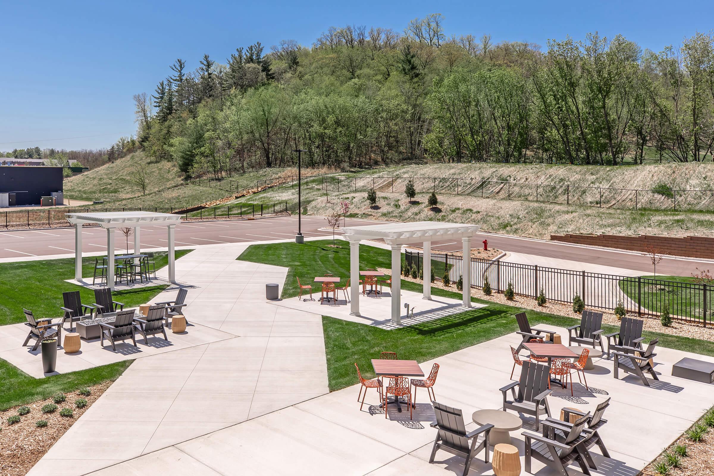 A spacious outdoor area featuring paved walkways, green grass, and several seating arrangements under white pergolas. Surrounding trees and a hillside provide a natural backdrop, while a road runs along the perimeter. The scene is bright and inviting, ideal for relaxation or social gatherings.