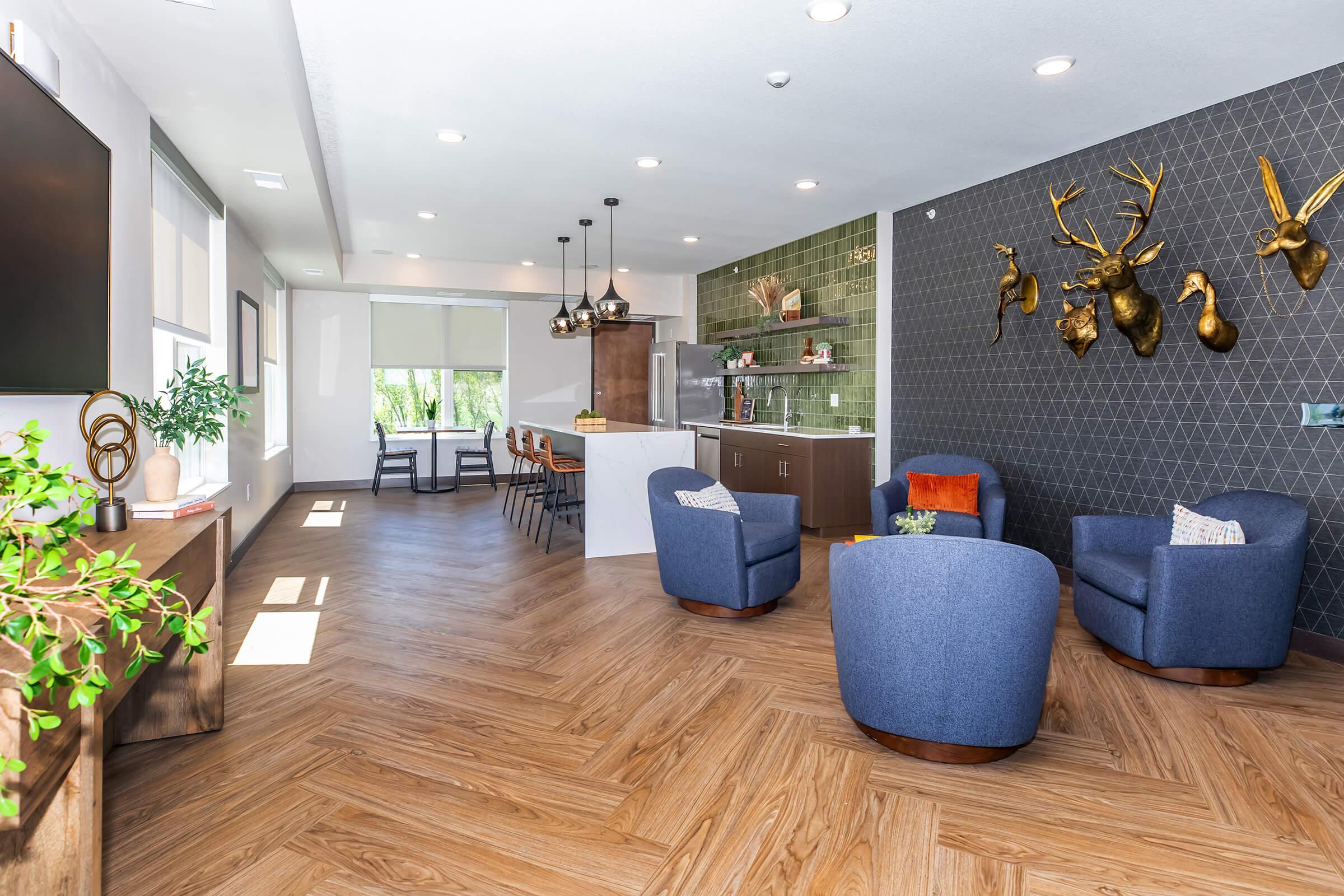 A modern lounge area featuring two blue armchairs, a wooden TV console, and a kitchenette with bar seating. The walls are decorated with geometric tiles and mounted deer head displays, while large windows allow natural light to fill the space. The flooring is a warm wood pattern, creating a welcoming atmosphere.