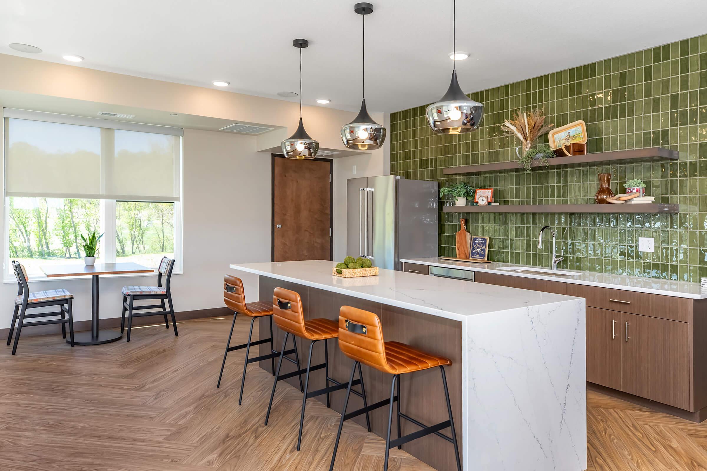 Modern kitchen featuring a large white island with brown bar stools, sleek cabinetry, and pendant lighting. The back wall is adorned with green tiles, and there's a dining area with a wooden table and black chairs near a window that overlooks greenery outside.