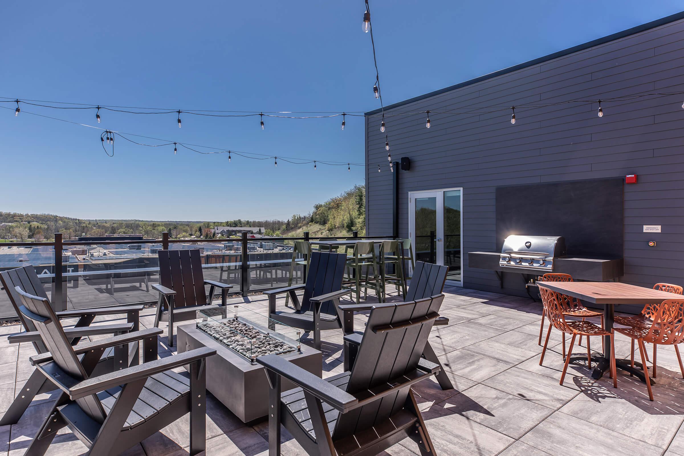 Rooftop patio featuring modern outdoor furniture, including black wooden chairs, a circular table with orange seating, and a gas grill. String lights hang overhead against a clear blue sky, with a scenic view of green hills in the background. A fire pit is centered among the seating area on the tiled floor.