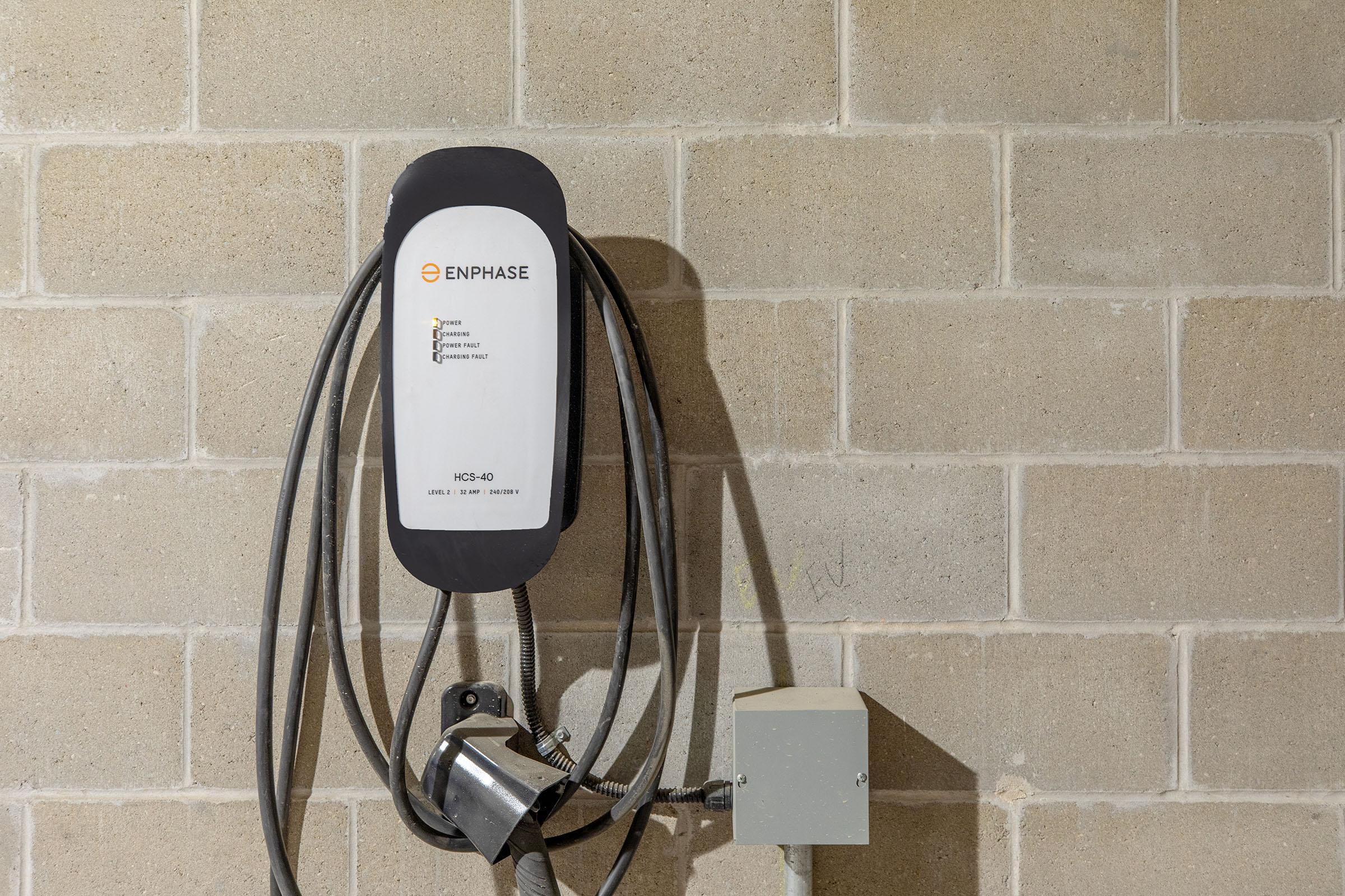 An Enphase electric vehicle charger mounted on a concrete wall, with two charging cables hanging down. A small electrical box is located below the charger. The setting appears to be a garage or indoor charging station.