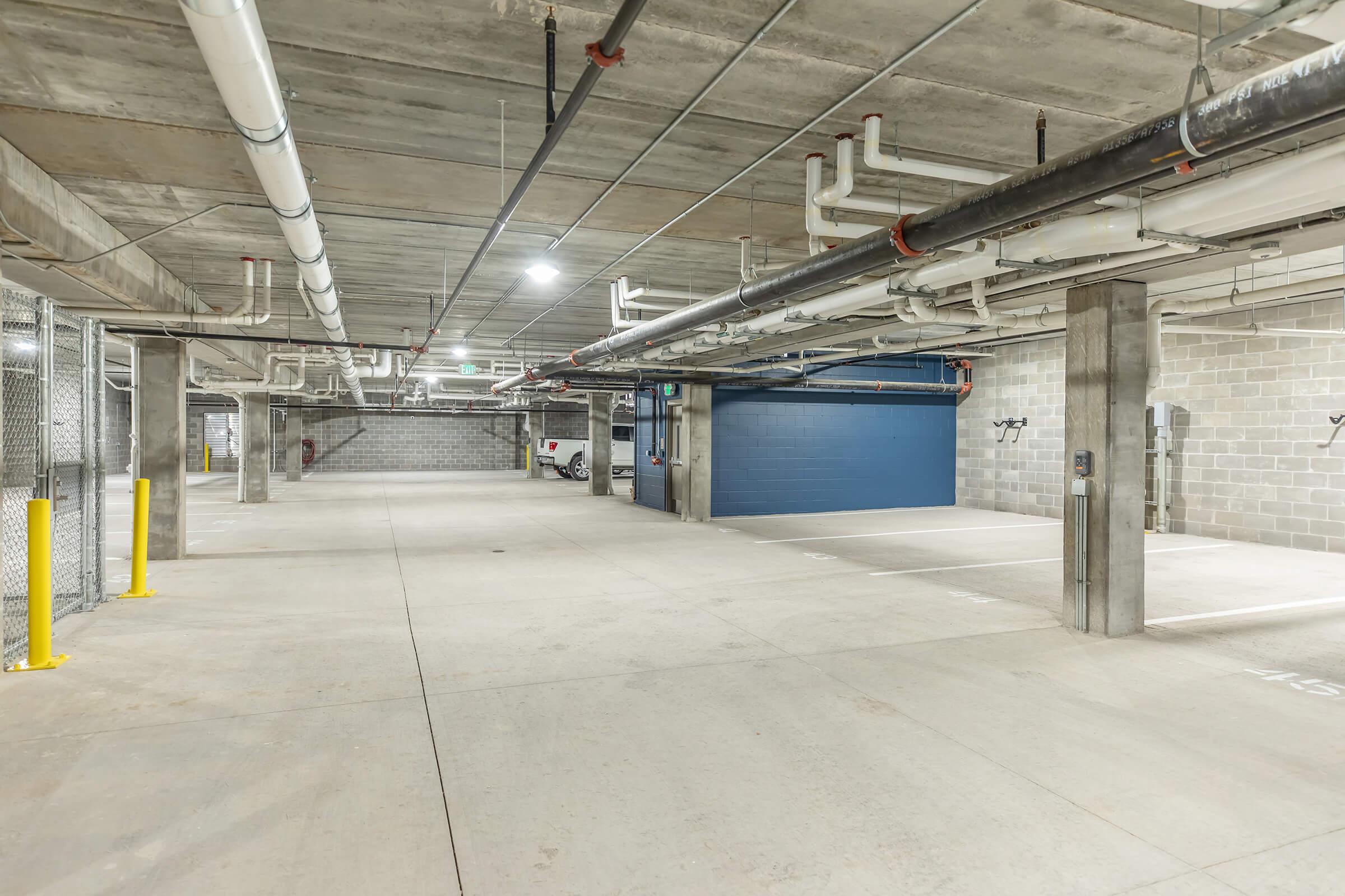 A spacious underground parking garage with concrete flooring, exposed pipes running along the ceiling, and bright overhead lights. Empty parking spaces are visible, along with a blue garage door at one end and sections of chain-link fencing in the background.