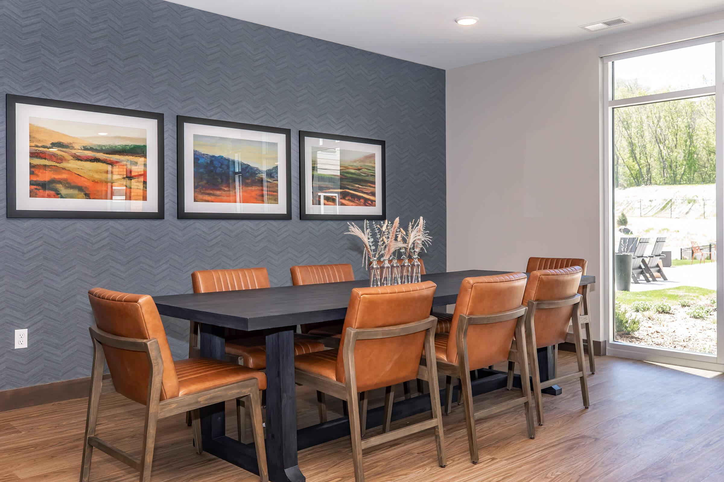 A modern dining room featuring a large wooden table surrounded by eight leather chairs. Three framed landscape artworks hang on the gray patterned wall. Natural light enters through a sliding glass door, which opens to an outdoor area with greenery and seating.