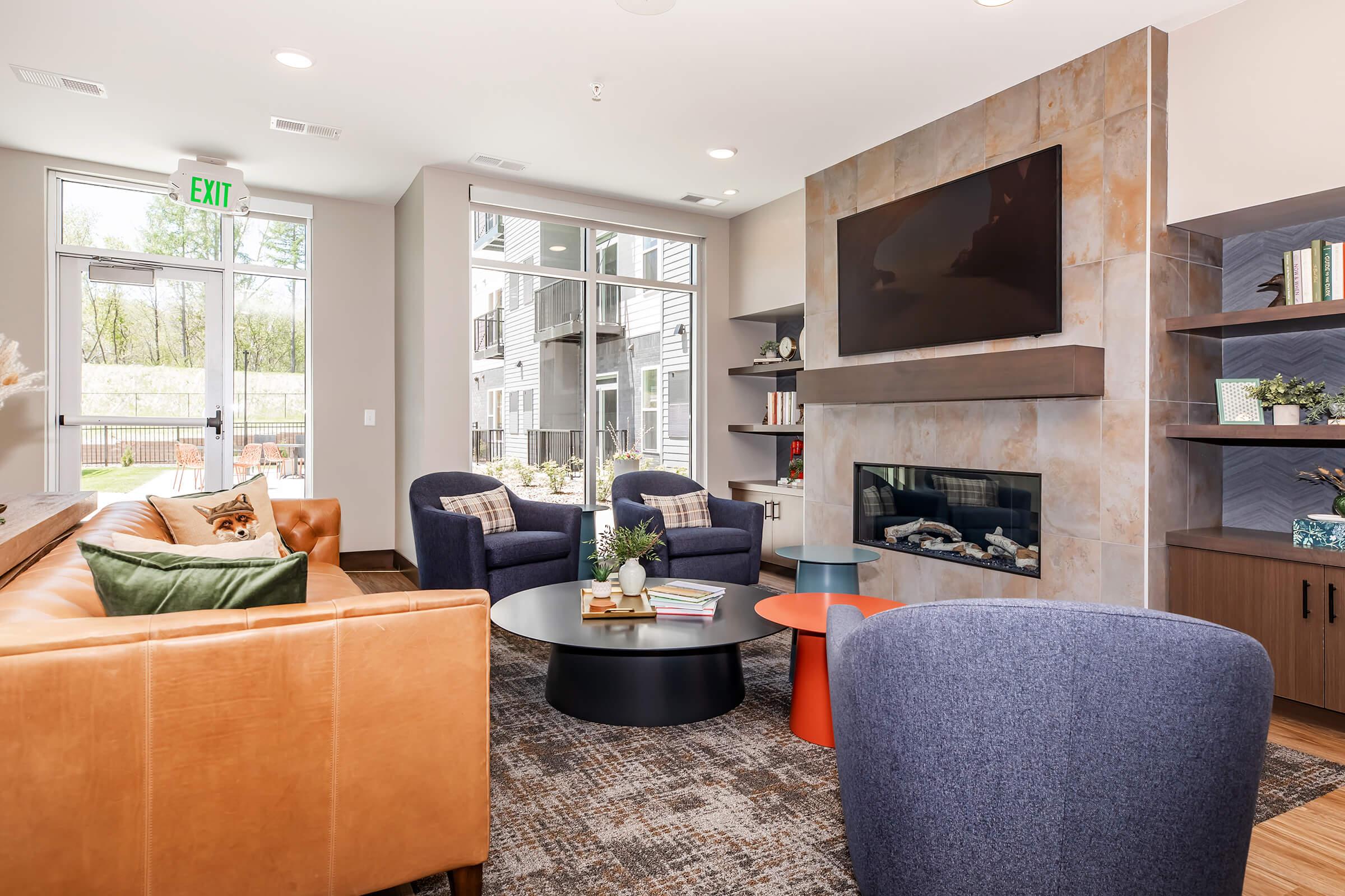 Cozy modern living space featuring a tan leather sofa, two blue armchairs, and a round coffee table. A mounted flat-screen TV is above a stylish fireplace, with built-in shelves displaying decorative items. Large windows let in natural light, and there are outdoor views of greenery.