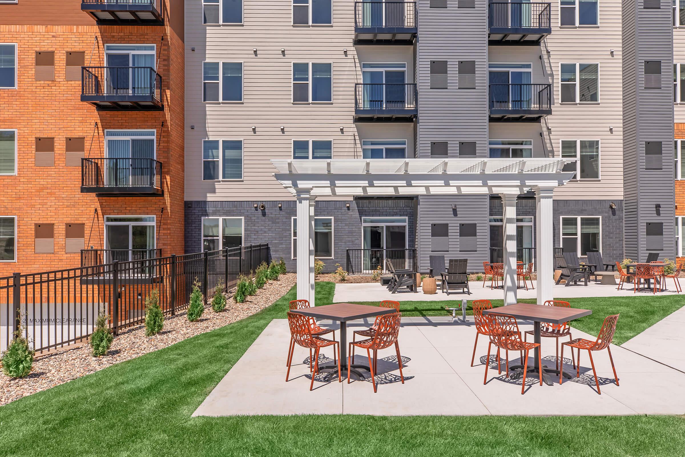 Outdoor patio area featuring modern furniture with tables and chairs, surrounded by green grass. In the background, there are multi-story residential buildings with balconies and a decorative pergola. The space is designed for relaxation and social gatherings.