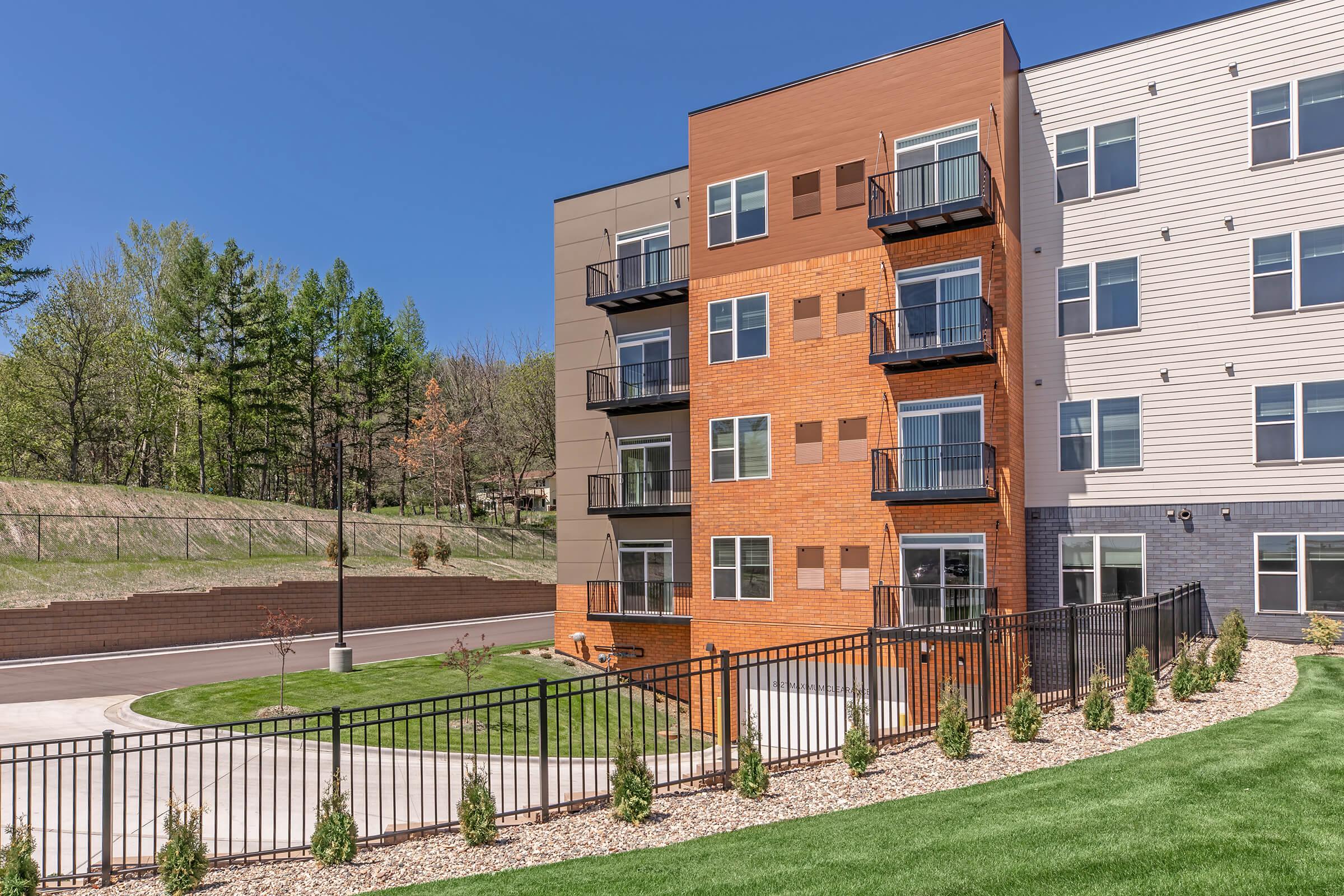 Modern apartment building featuring a mix of brown and gray exterior with balconies, surrounded by green grass and small shrubs. A well-maintained landscape and a fenced area can be seen in the foreground, with trees in the background under a clear blue sky.