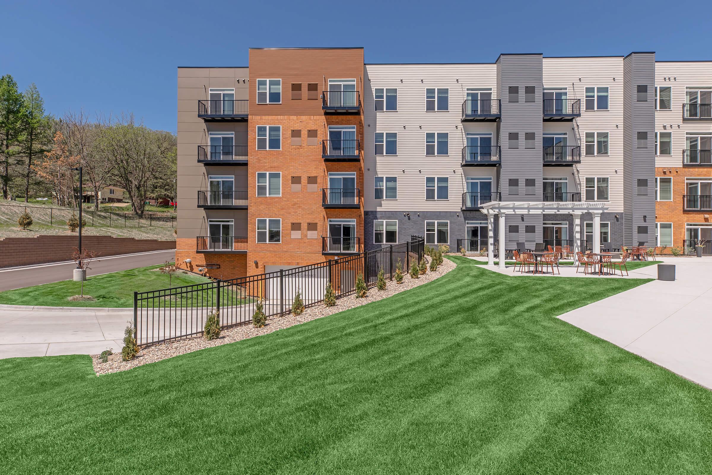Modern apartment building with a mix of orange and gray exterior, featuring balconies on upper floors. Well-maintained green lawn and landscaping in the foreground, with a patio area that includes seating and a pergola. Clear blue sky and trees in the background provide a pleasant setting.