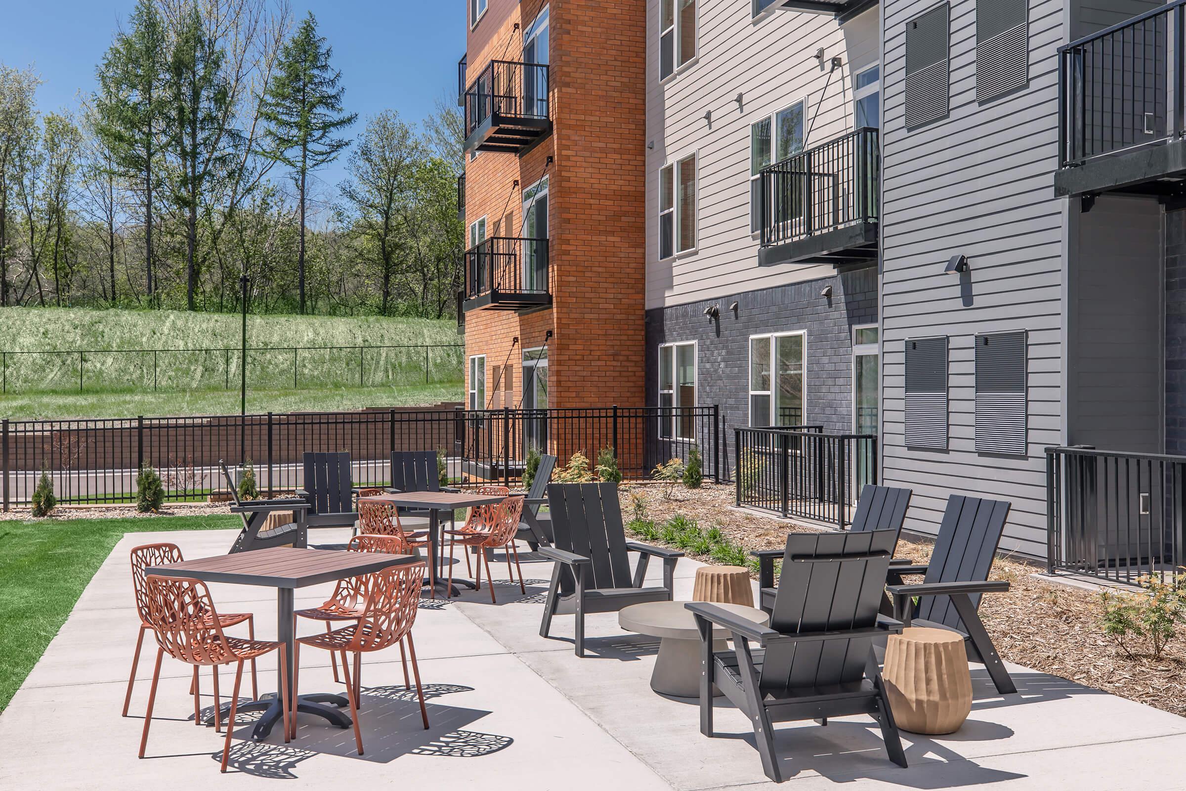 Outdoor patio area with various seating options, including wooden and metal chairs and tables. Modern apartment buildings in the background, surrounded by green grass and trees. A fire pit and planters add to the inviting atmosphere for relaxation and social gatherings. Clear blue sky enhances the overall ambiance.