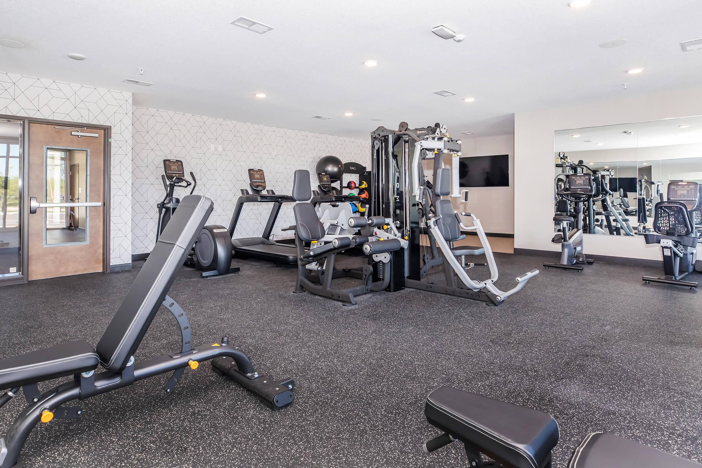 A modern gym interior featuring various exercise equipment including weight benches, cardio machines, and a multi-station workout machine. The space has a clean, bright design with large mirrors and adequate lighting, suitable for various fitness activities.