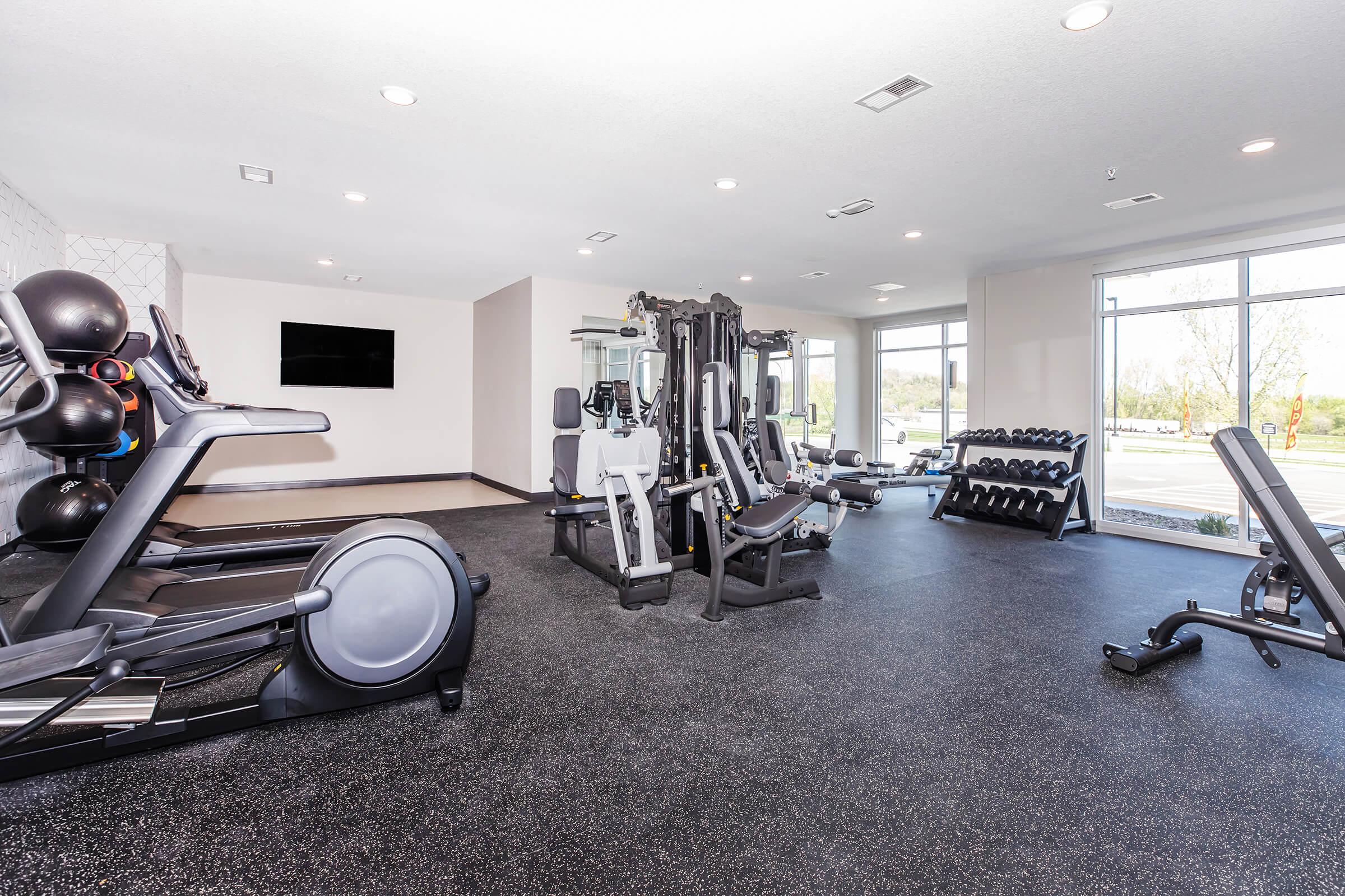 A modern gym interior featuring cardio machines, weight training equipment, and free weights. The space includes a large window allowing natural light and a wall-mounted TV. The flooring is dark and rubberized, providing a safe and comfortable workout environment.
