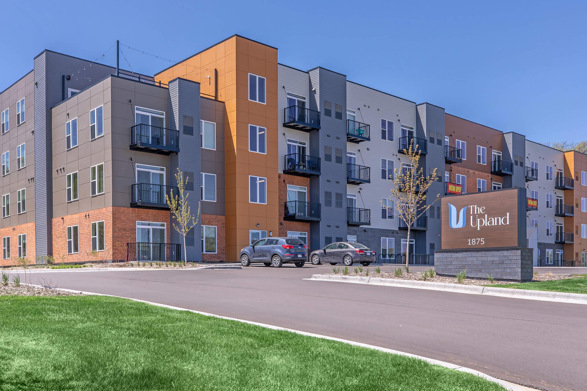 Modern apartment complex with a mix of gray and orange facade. Balconies on several units. In front, a landscaped area with a sign reading "The Upland" and the address "1875." Paved parking area with a few cars visible, surrounded by green grass.