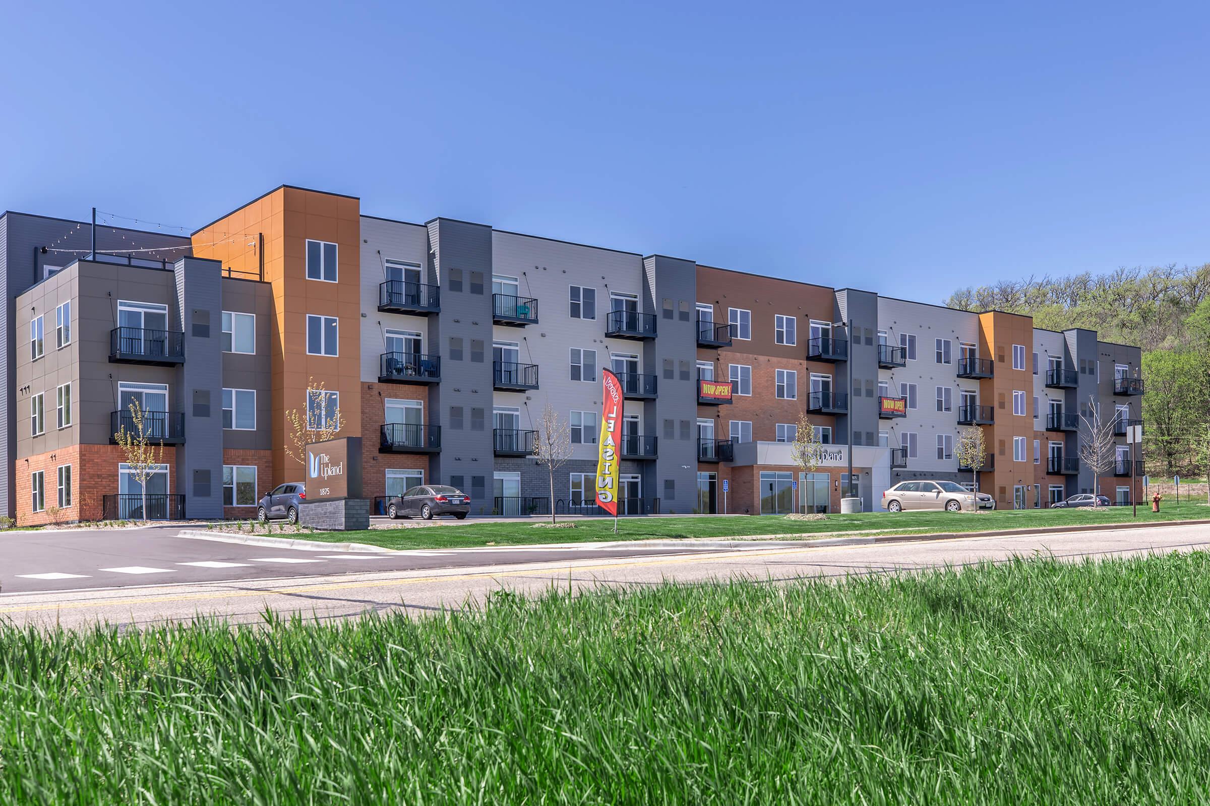 Modern apartment building with a mix of orange, gray, and beige facades. Several balconies are visible, and there are cars parked in front. The foreground features green grass, and a clear blue sky is in the background, indicating a bright, sunny day.