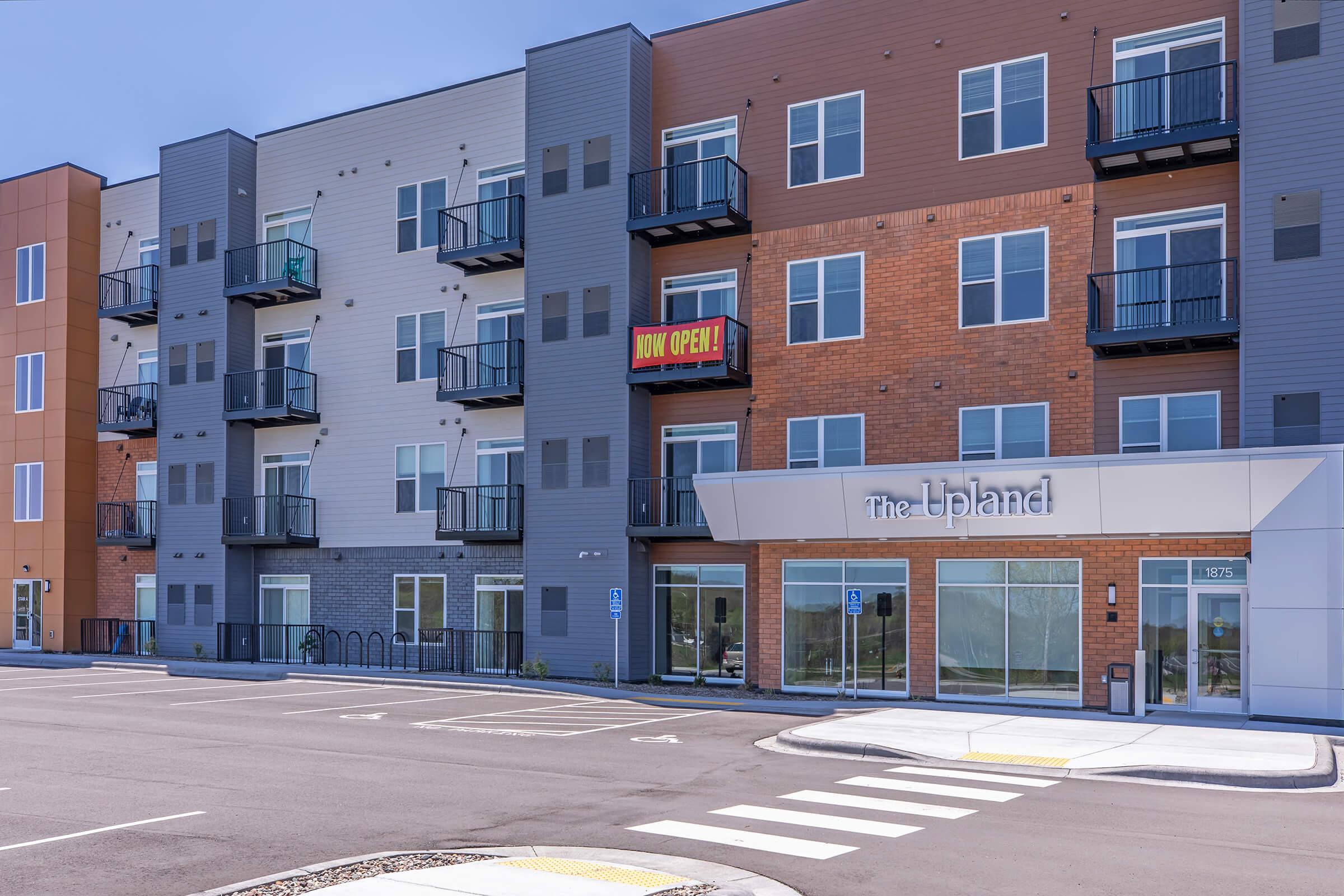 Modern apartment building with a mix of brick and gray siding. The entrance features large windows and a sign that reads "Now Open!" There are balconies on the upper floors, and the surrounding area is paved with parking spots and bike racks. Clear blue sky overhead.