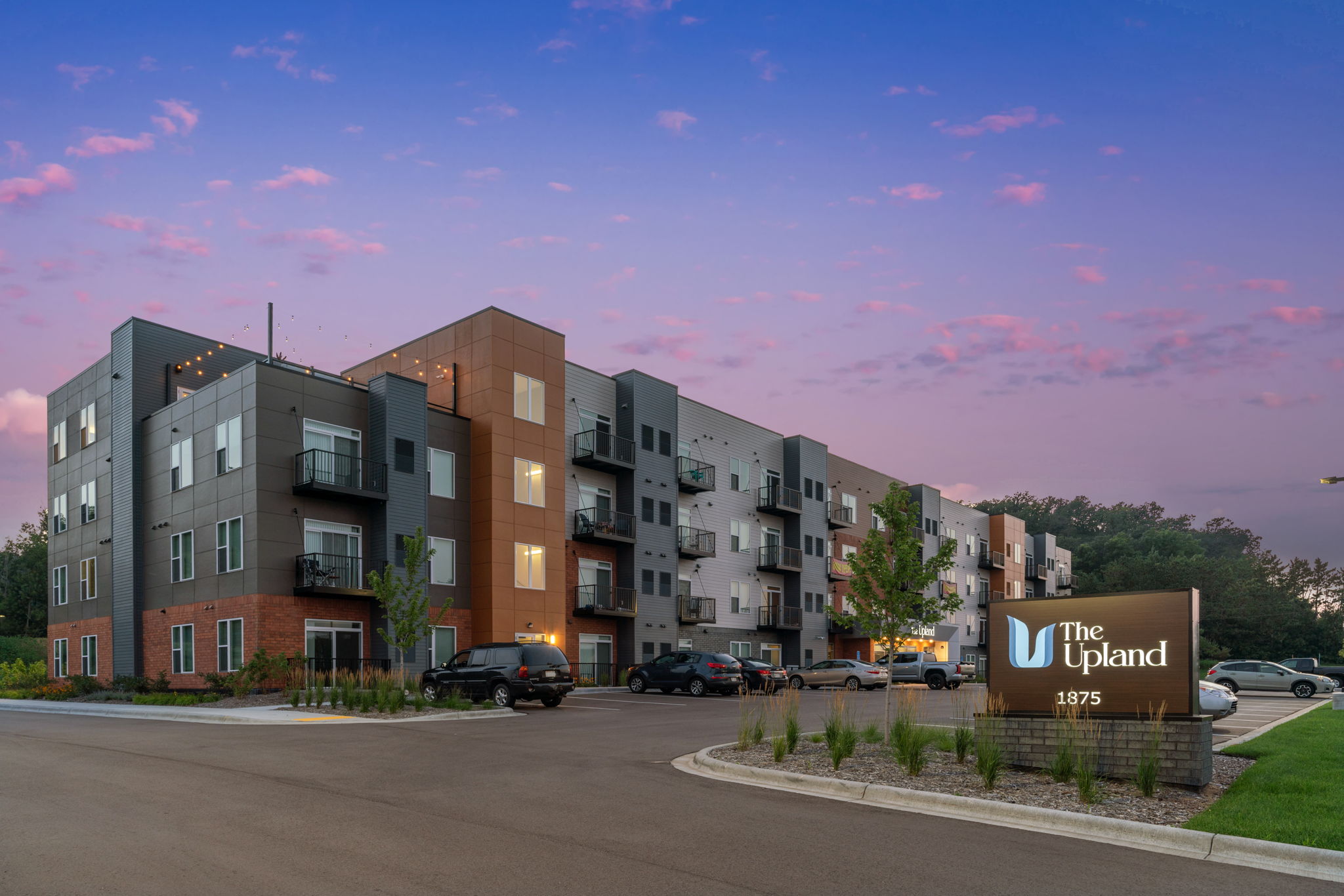 Modern apartment complex with a stylish facade, featuring a mix of brick and metal siding. The Upland sign is prominently displayed in the foreground, surrounded by landscaped greenery and parking spaces. The sky is colorful with pink and purple hues, suggesting either dawn or dusk.