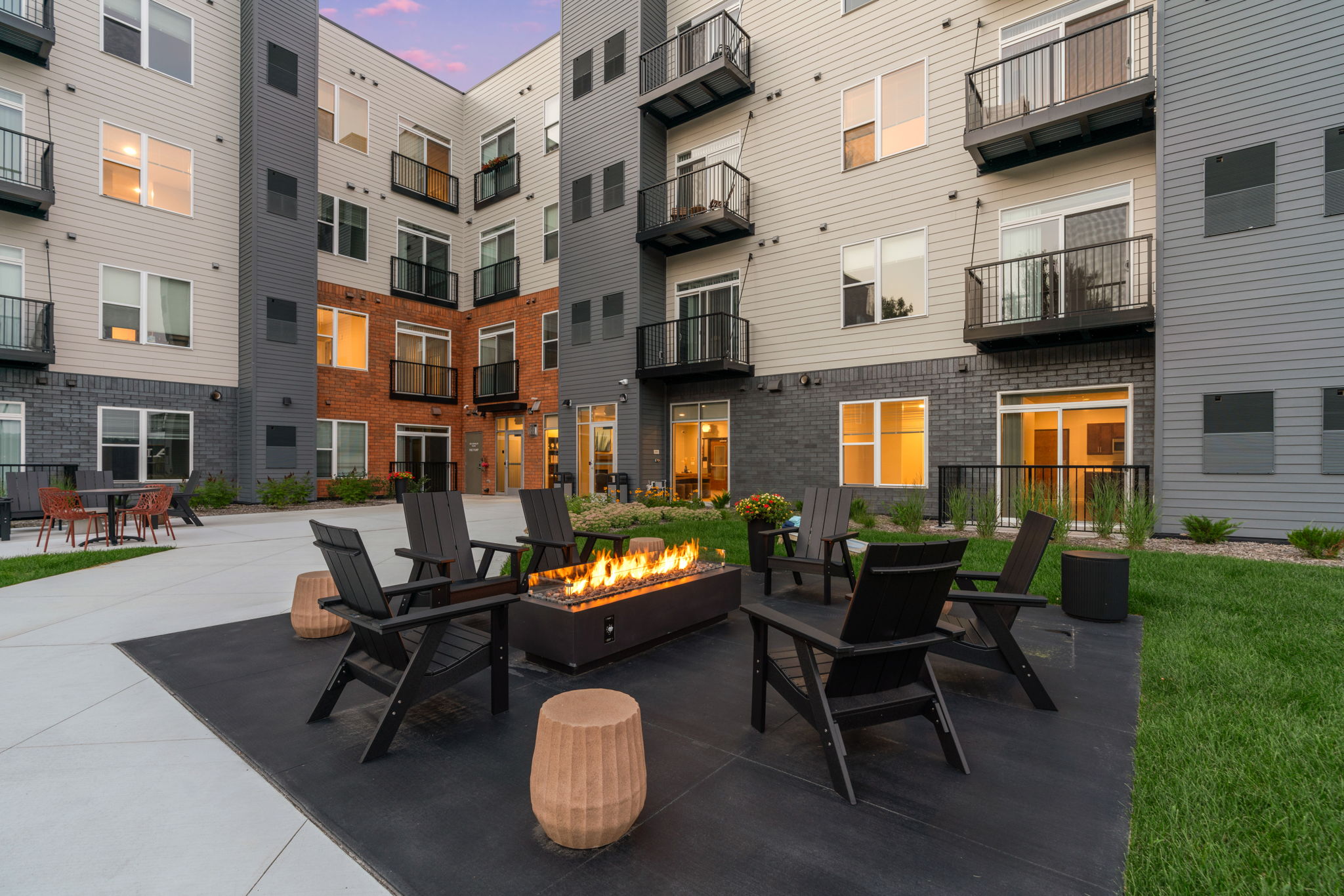 A modern apartment complex courtyard featuring multiple buildings with balconies. In the center, there is a stylish fire pit surrounded by black patio chairs. The area is well landscaped with grass and flower beds, and a few outdoor tables in the background. Warm evening light glows from the windows.