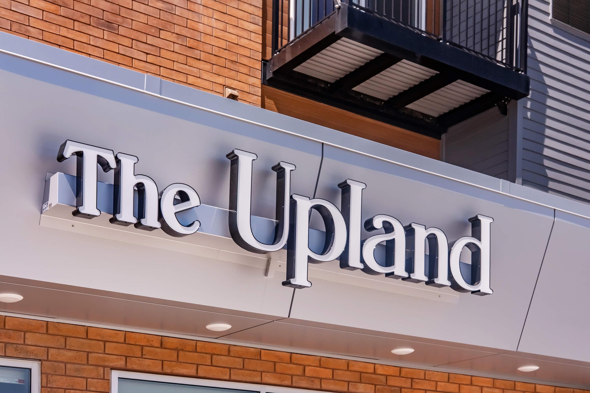 Signage for "The Upland" displayed prominently on a building exterior, featuring bold, stylized lettering against a brick wall. The sign is well-lit and clearly visible, highlighting the establishment's name.