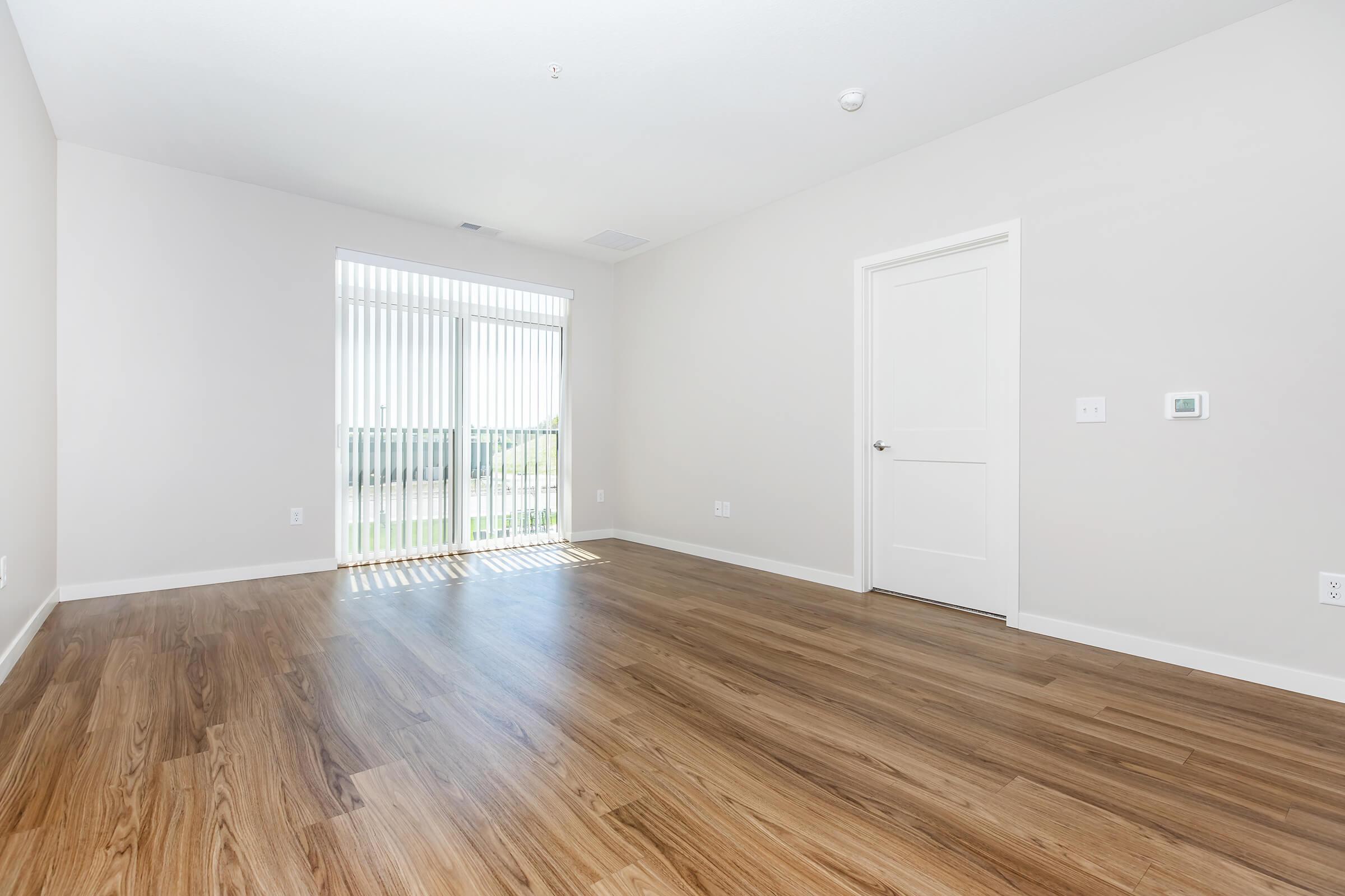 A vacant room with light hardwood flooring, featuring a large window with vertical blinds that lets in natural light. The walls are painted a light color, and there is a closed door on the right side of the image. The overall ambiance is bright and spacious.