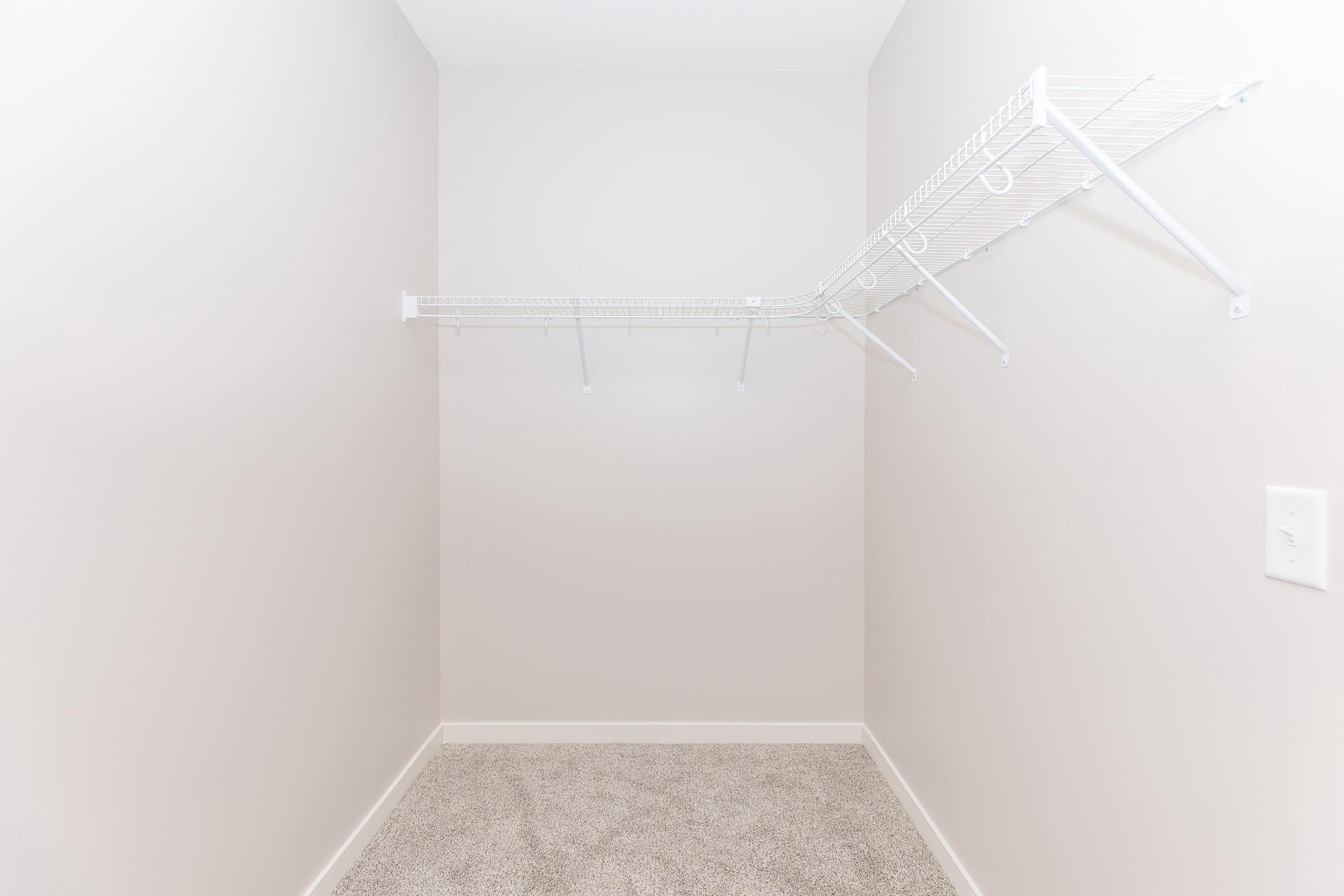 A small empty closet with light beige walls and a carpeted floor. There is a wire shelving unit mounted on one wall, with several hooks and no items displayed, creating a spacious and minimalistic appearance.