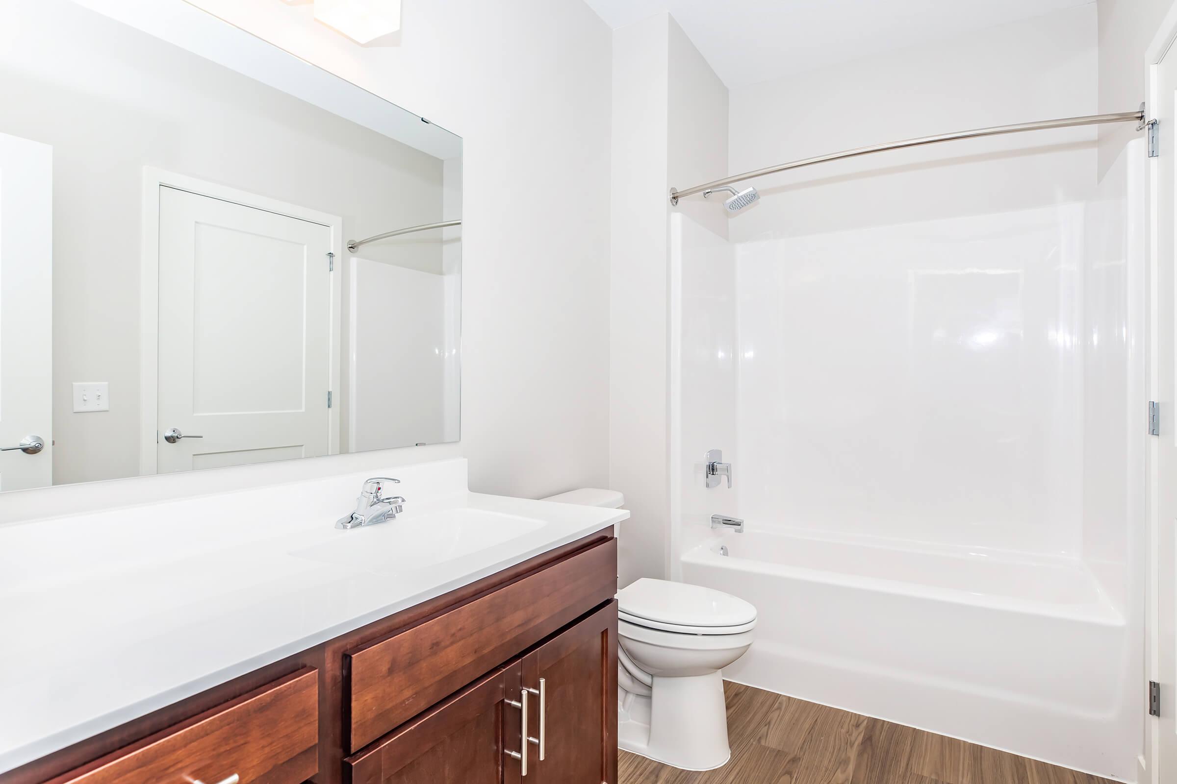 A clean, modern bathroom featuring a white bathtub and shower with a glass door, a large mirror above a sleek vanity with a brown wood finish, a white toilet, and neutral-colored walls and flooring. The space is well-lit and organized, creating a fresh and inviting atmosphere.