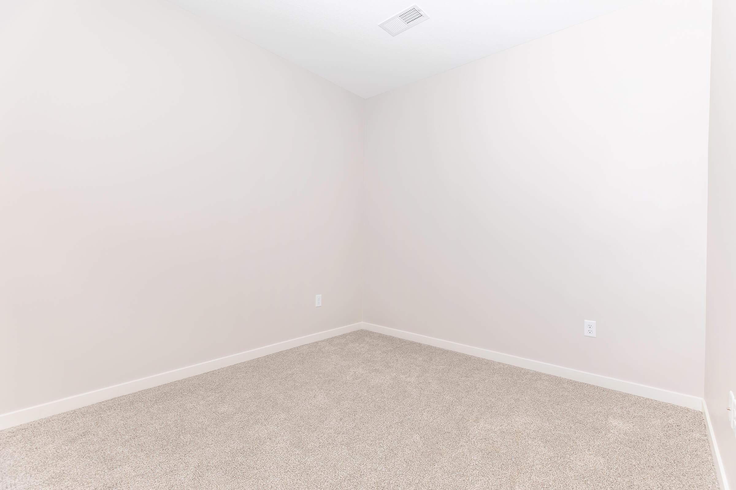 A spacious, empty room with light beige walls and soft beige carpet. The room features a single light fixture on the ceiling and a vent, with no furniture or decorations. Natural light illuminates the space, giving it a bright and airy feel.