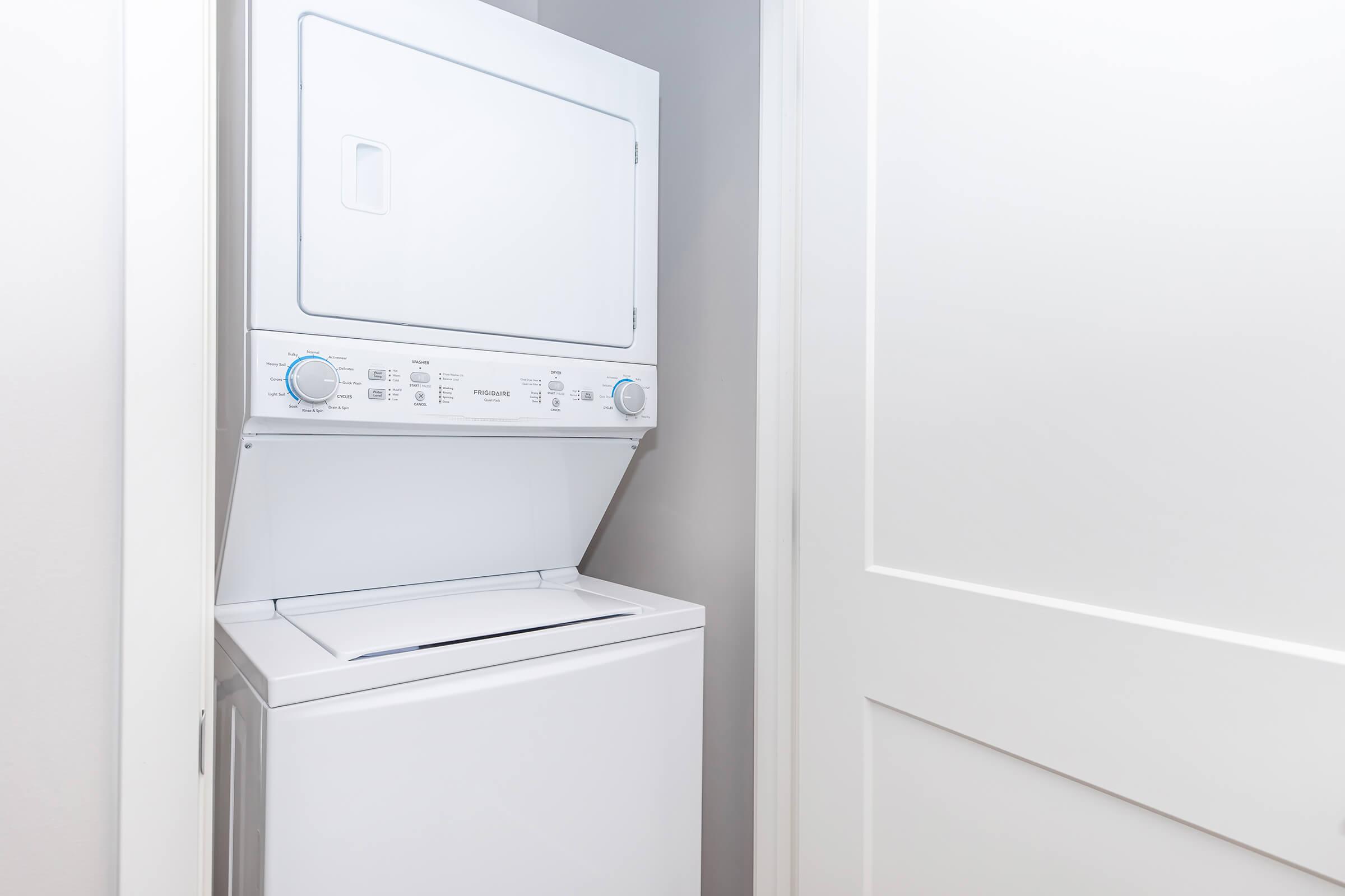 A stackable washer and dryer unit placed against a light-colored wall, with a closed door beside it. The appliance features control dials and a front-loading dryer on top. The setting is clean and modern, reflecting a functional laundry area in a home.