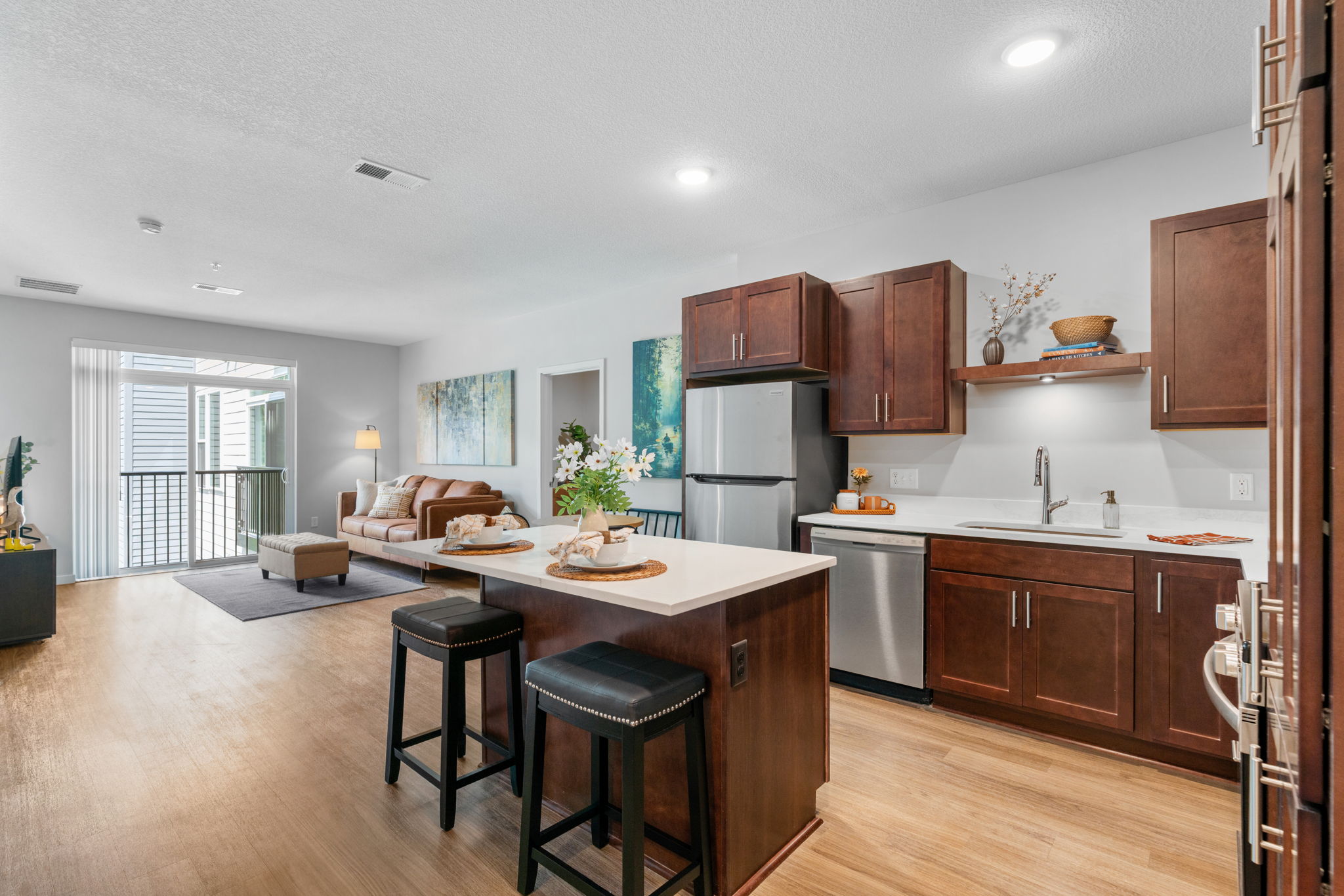 Modern apartment interior featuring a kitchen with dark wood cabinets and stainless steel appliances. A central island with bar stools is in front, while a cozy living area with a sofa and artistic wall decor is visible in the background. Natural light enters through sliding glass doors.