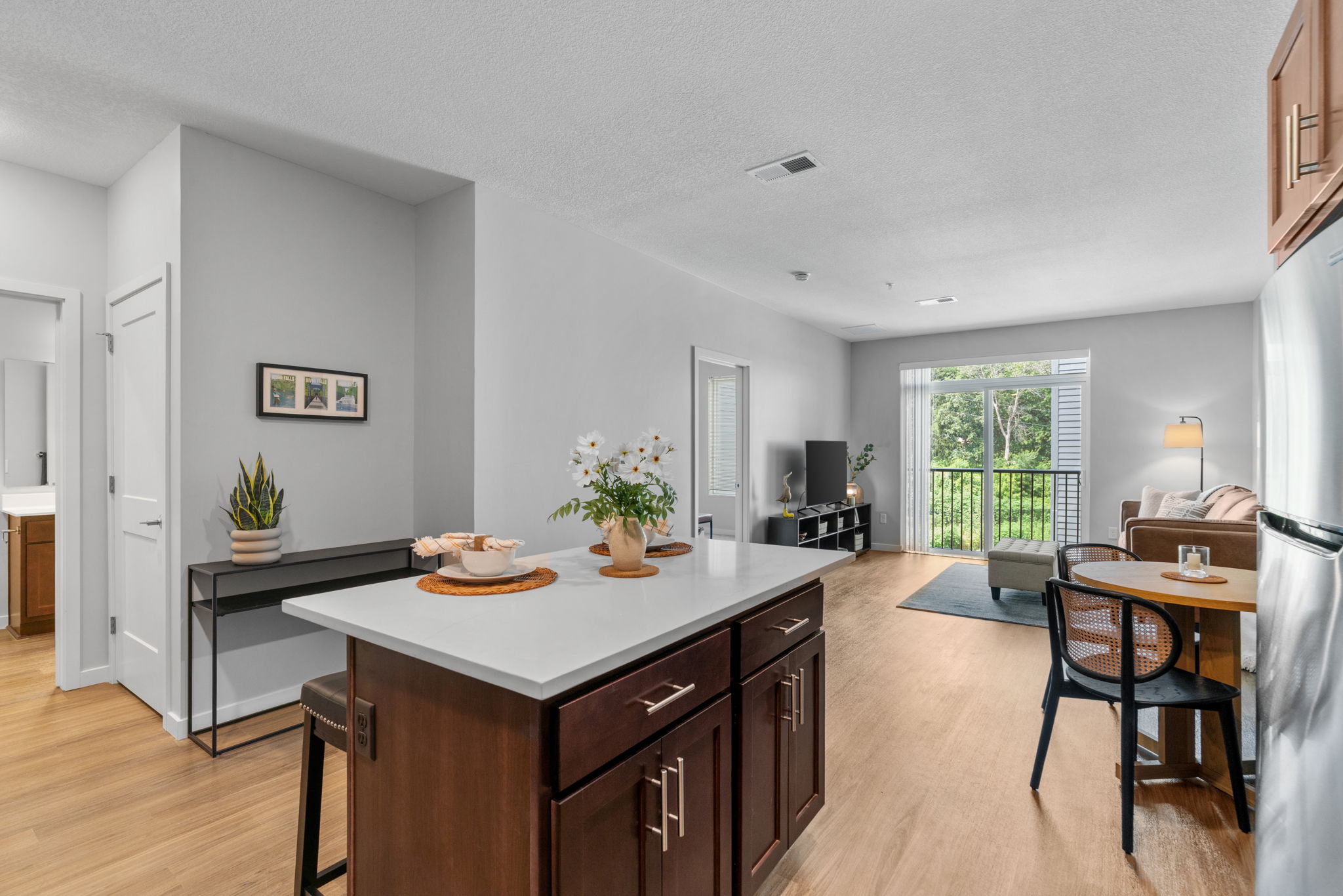A modern apartment interior featuring a kitchen island with a vase of flowers, small dining table, and living area with a couch and TV. Large windows allow natural light to fill the space, creating a bright and inviting atmosphere. Neutral colors and wooden flooring enhance the contemporary design.