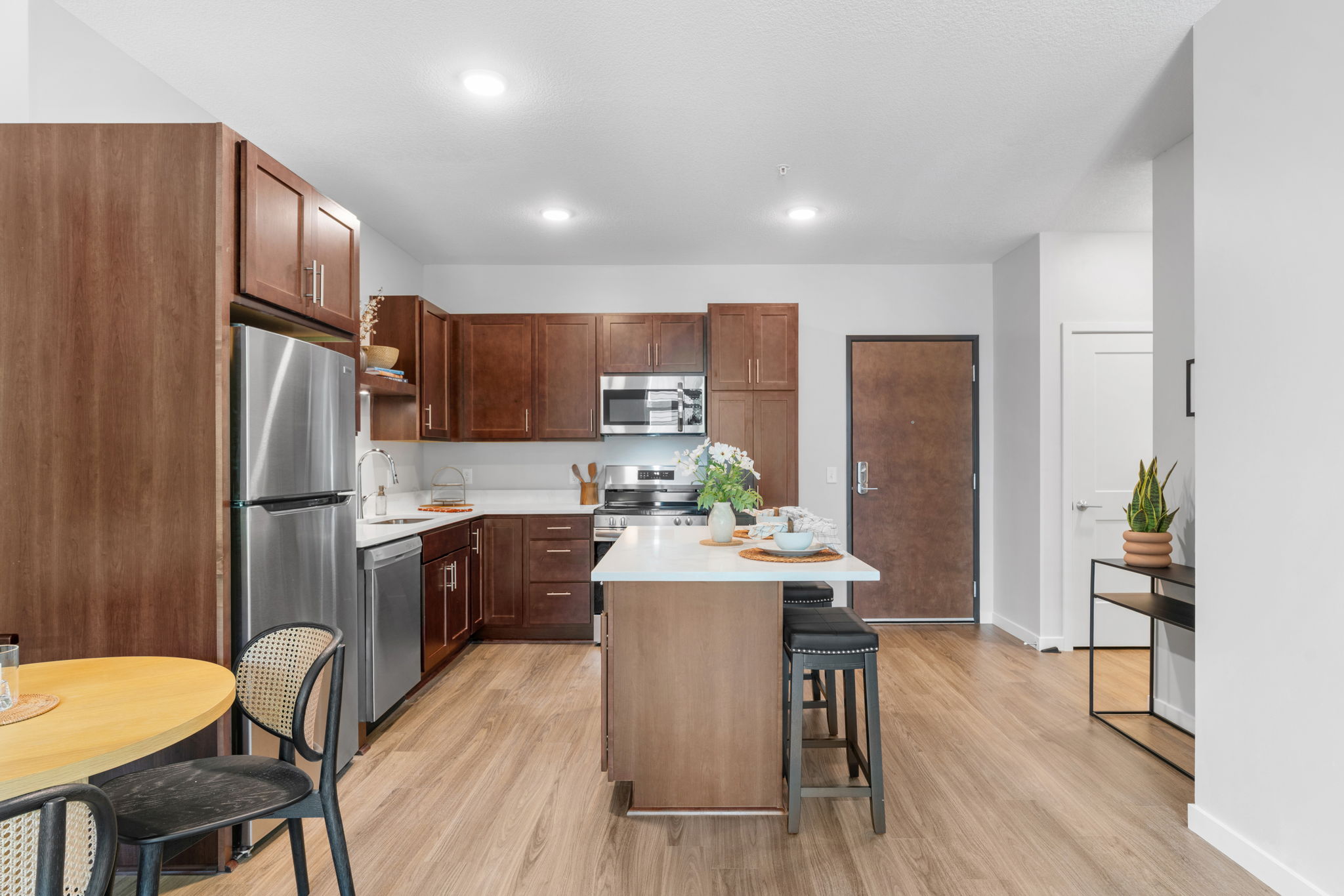 Modern kitchen with wooden cabinets, stainless steel appliances, and an island with seating. A small dining table is set for two. The space features wood-like flooring and minimalistic decor, with a potted plant near the entrance. Natural light brightens the area, creating an inviting atmosphere.
