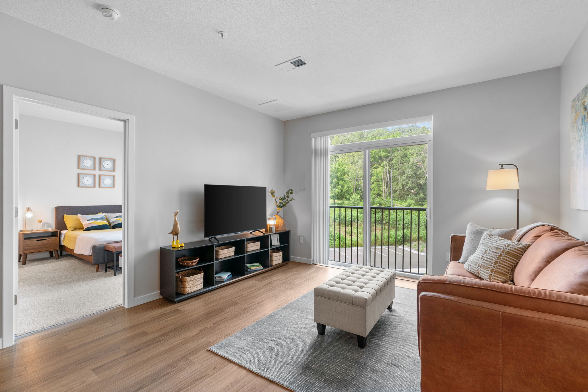 A cozy living room featuring a brown sofa, a footstool, and a TV stand with decorative items. Large windows provide natural light and a view of greenery outside. An open doorway leads to a bedroom, with colorful bedding visible. The space has a modern, inviting feel with neutral colors and warm wood flooring.