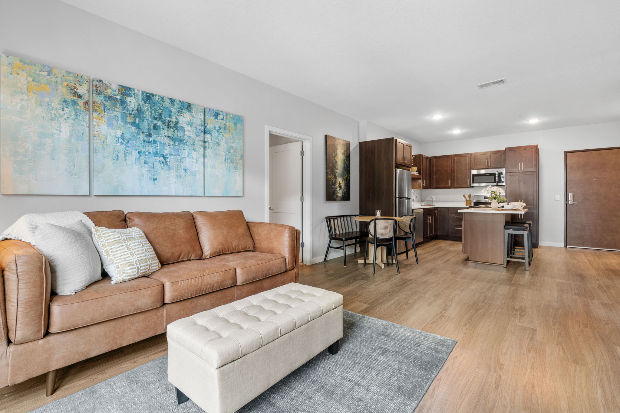 A modern living room with a brown leather sofa, a light-colored ottoman, and a gray area rug. Abstract art hangs on the wall above the couch. In the background, a kitchen features dark wooden cabinets and stainless steel appliances, with a dining area that includes black chairs and a table. Natural light fills the space.