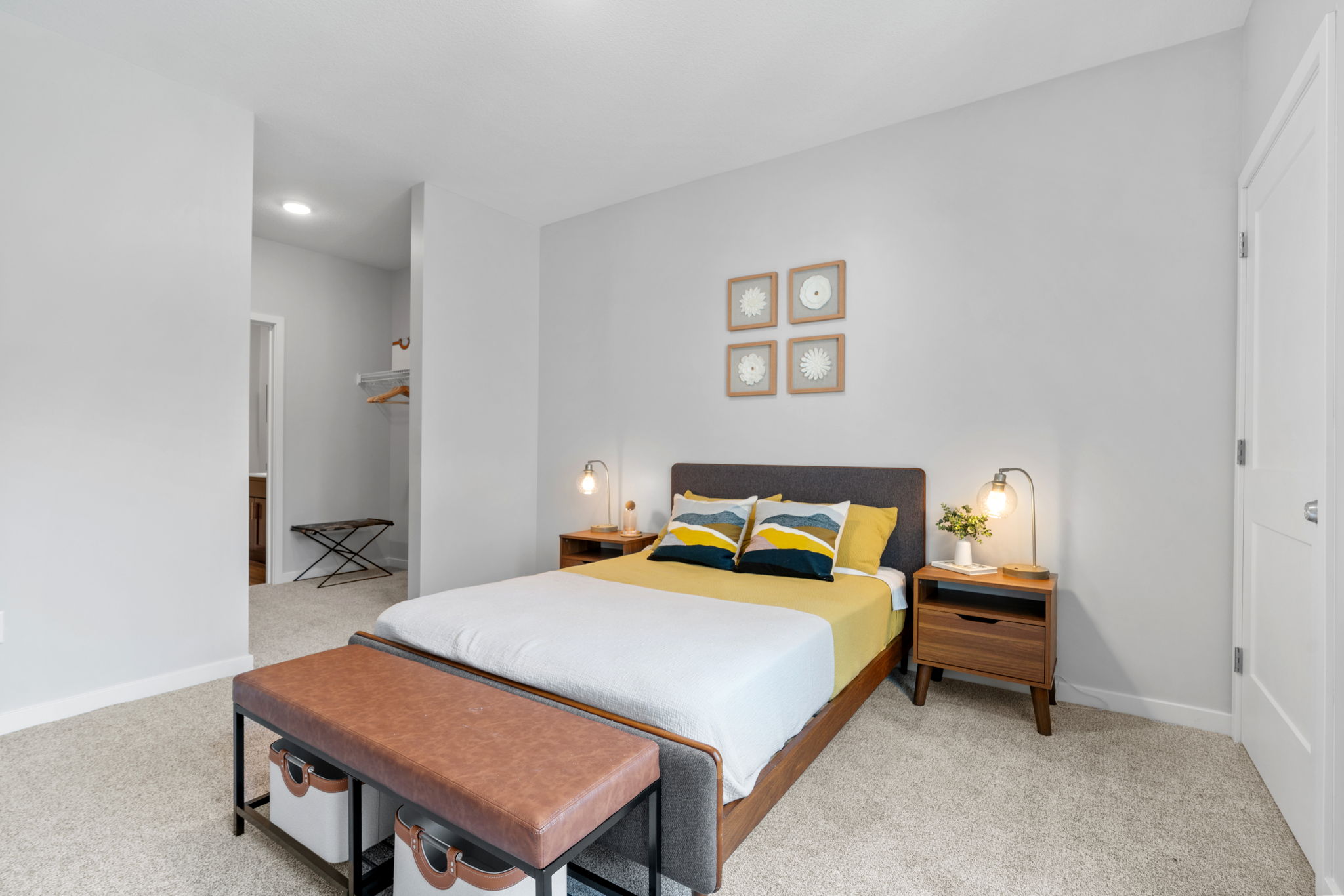 A neatly arranged bedroom featuring a bed with yellow and blue pillows, a brown bench at the foot, two wooden nightstands with lamps, and light gray walls. The room has a cozy carpet and an open closet area, creating a modern and inviting atmosphere.